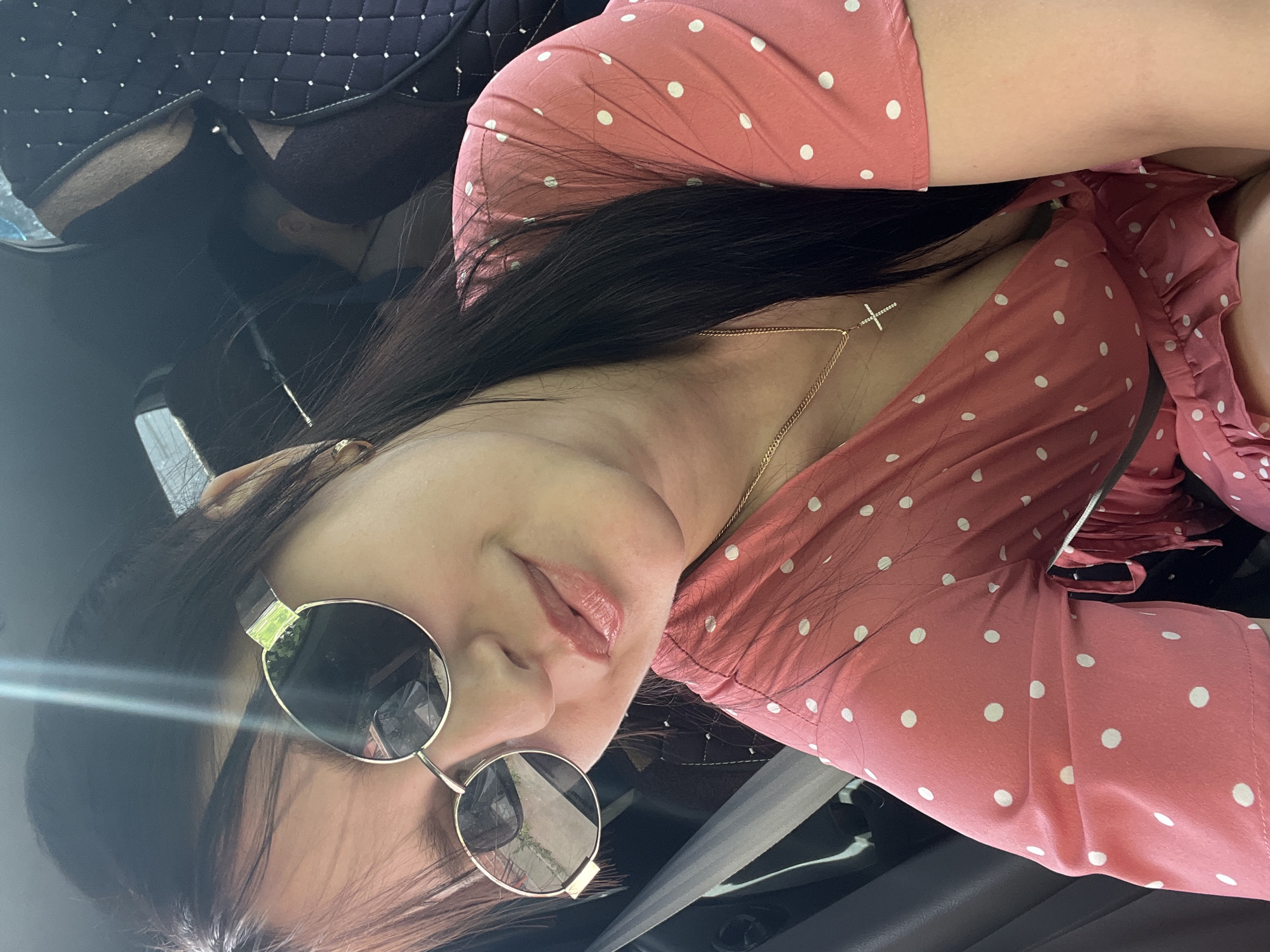 The image is a close-up, selfie-style shot of a woman wearing round sunglasses and a pink polka-dot dress, taken from a slightly angled perspective inside a car. She has dark hair and is wearing a delicate gold chain necklace with a cross pendant. The background is the interior of the car, including the seat and a partially visible headrest cover with a quilted pattern. The location is not evident within the image; it is impossible to verify the location to be Chaikovsky, Russia.