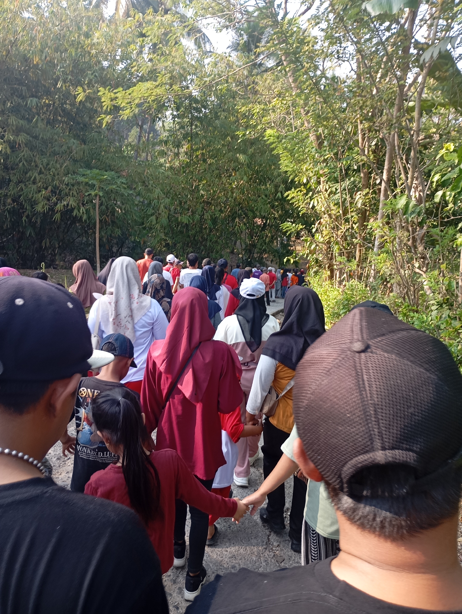 A group of people are walking along a path lined with trees and bamboo in what appears to be an outdoor recreational area. The path is made of gravel. In the foreground, the backs of two individuals are visible, one wearing a black baseball cap and the other a dark brown baseball cap. The group ahead consists of at least a dozen individuals, predominantly wearing headscarves, with clothing in various colors including red, white, beige, and blue. The sun is casting shadows, indicating it is likely daytime. The environment is lush with green foliage. The context of this image is a gathering or procession taking place in Ketepung, Indonesia.
