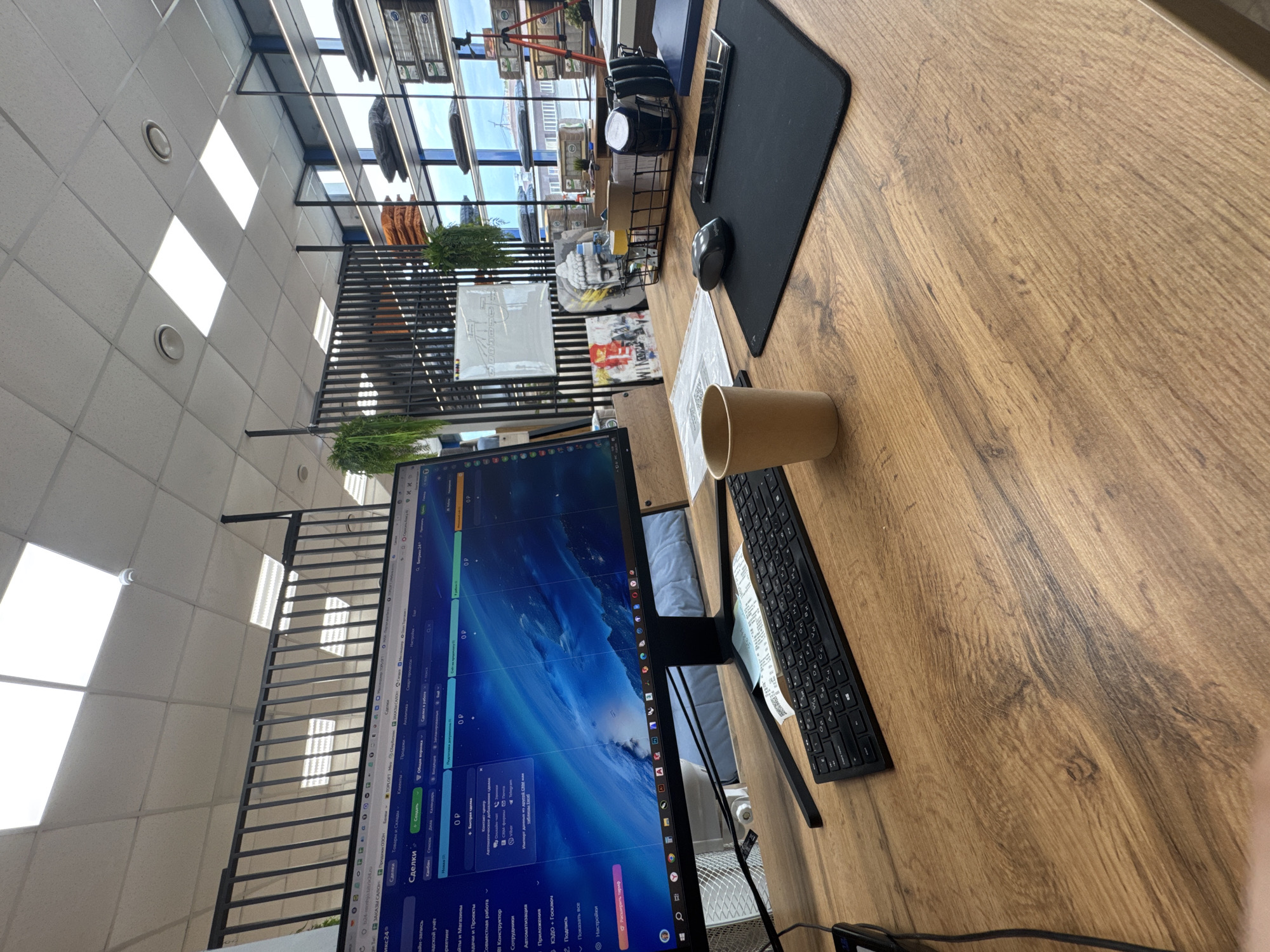 The image captures a modern office workstation from a high, tilted perspective, showcasing a wooden desk and surrounding environment. In the foreground, a computer monitor displays a software interface, likely a CRM or project management system like Bitrix24. Visible text on the screen includes categories such as "Сделки" (Deals), "Товары и Склады" (Goods and Warehouses), "Клиенты" (Clients), "Продажи ОЗОН" (Ozon Sales), "TOPLOFT-M", and "Банки" (Banks). Other options like "Автоматизация" (Automation), "Приложения" (Applications), "КЭДО + Госключ" (EDM + Gosklyuch), and "Подпись" (Signature) are also seen, alongside "Bitrix24.ru".
A black keyboard is placed in front of the monitor, with a black mouse and mousepad to its right. A light brown paper cup rests on the desk. Further back, a desk organizer holds various office supplies, a mobile phone, and a small decorative statue. Several papers, including one with a QR code, are scattered on the desk.
The background features a grid-patterned office ceiling with bright rectangular light fixtures and circular vents. A large window provides natural light and a view of an external building or structure under a clear sky. A black metal slatted divider or railing, adorned with artificial greenery, separates the workstation from the background. The scene depicts a well-lit, functional workspace, likely during business hours.
