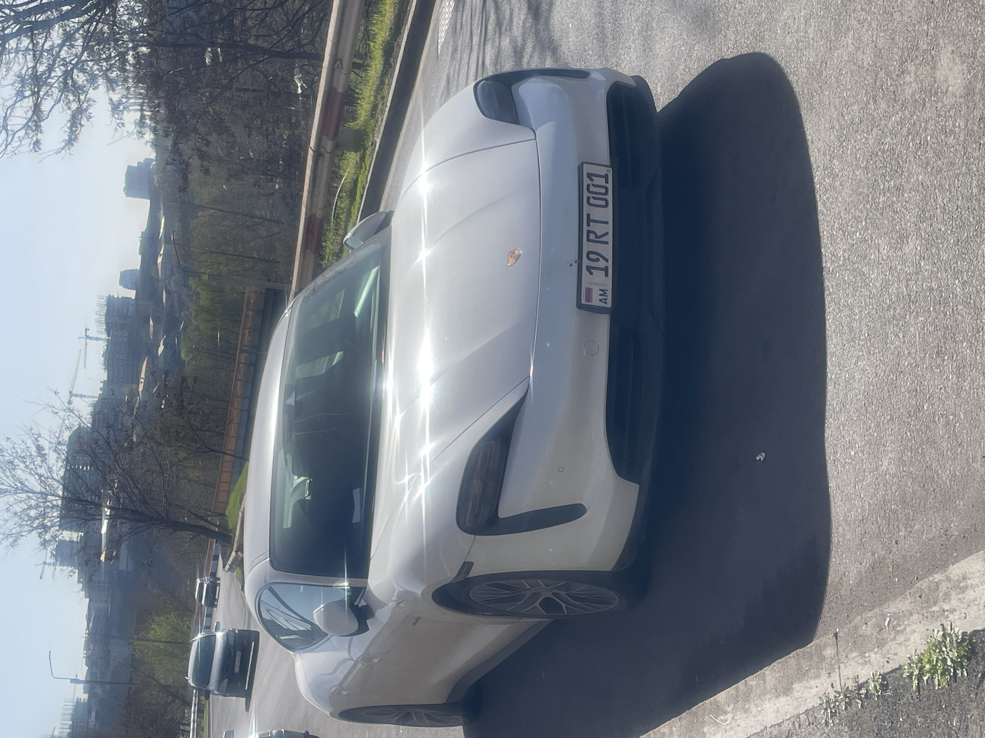 A white Porsche Taycan is parked on a street in Yerevan, Armenia. The license plate reads "19 RT 001" and the country code "AM" is visible. The photo is taken at street level. In the background, buildings and trees are visible, indicating an urban area. There are also other cars driving in the same street in the background.