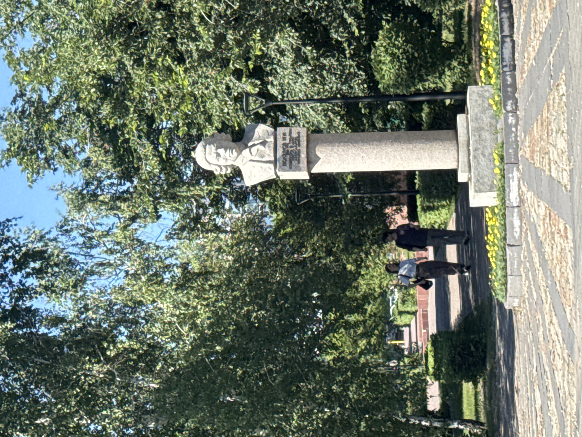 The image shows a monument in a park in Barnaul, Russia. The monument is a bust of a man on a pedestal. The man has curly hair and is wearing a suit. There are two people walking on a path behind the monument. The path is lined with trees and bushes. There are also some yellow flowers in the foreground. The sky is blue and there are some clouds.