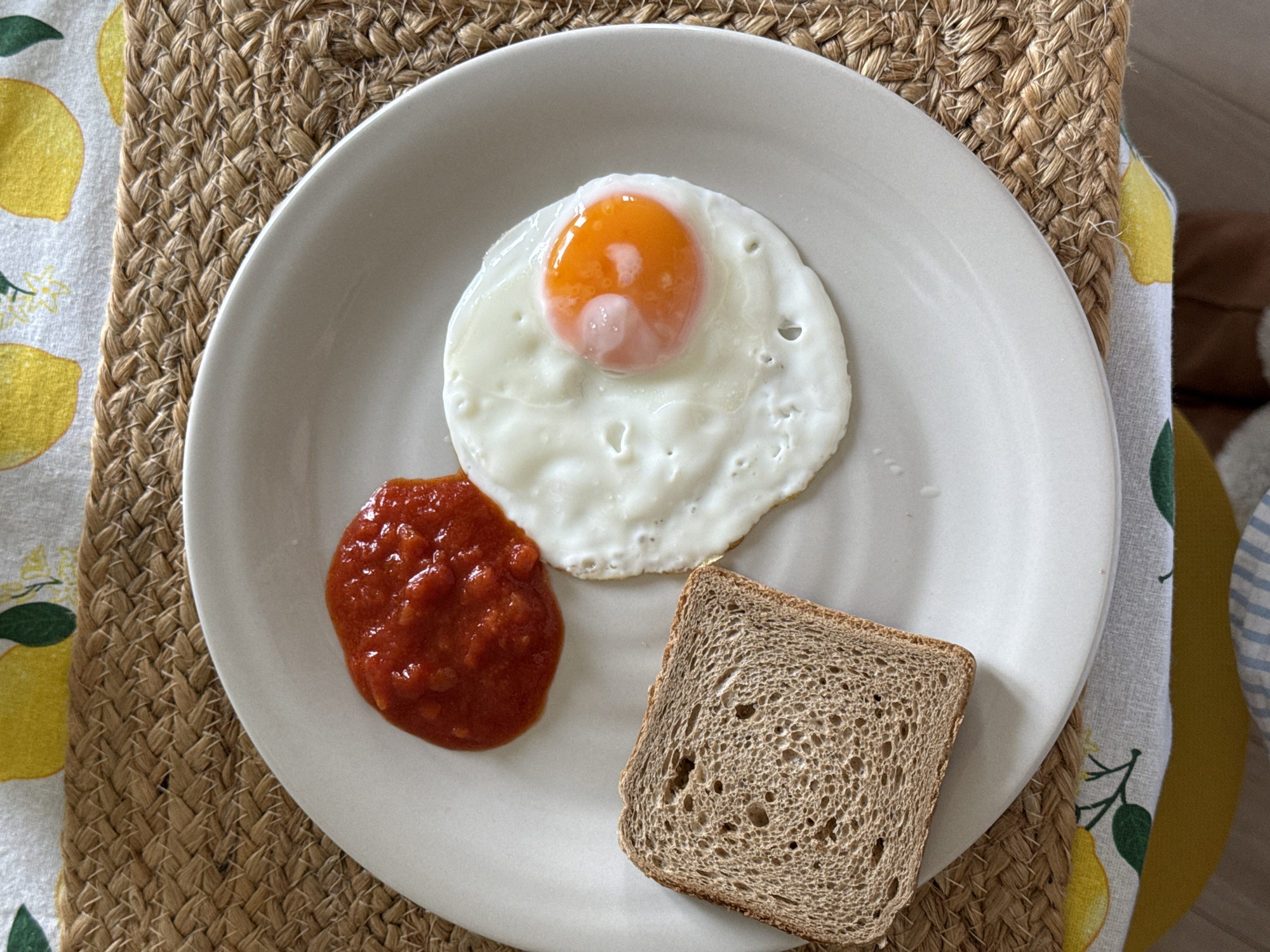A plate of food sits on a woven placemat, which is partially overlaid by a fabric with a lemon print. On the plate, there is a fried egg with a runny yolk, a dollop of red sauce, and a single slice of brown bread. The location is impossible to determine from the image.