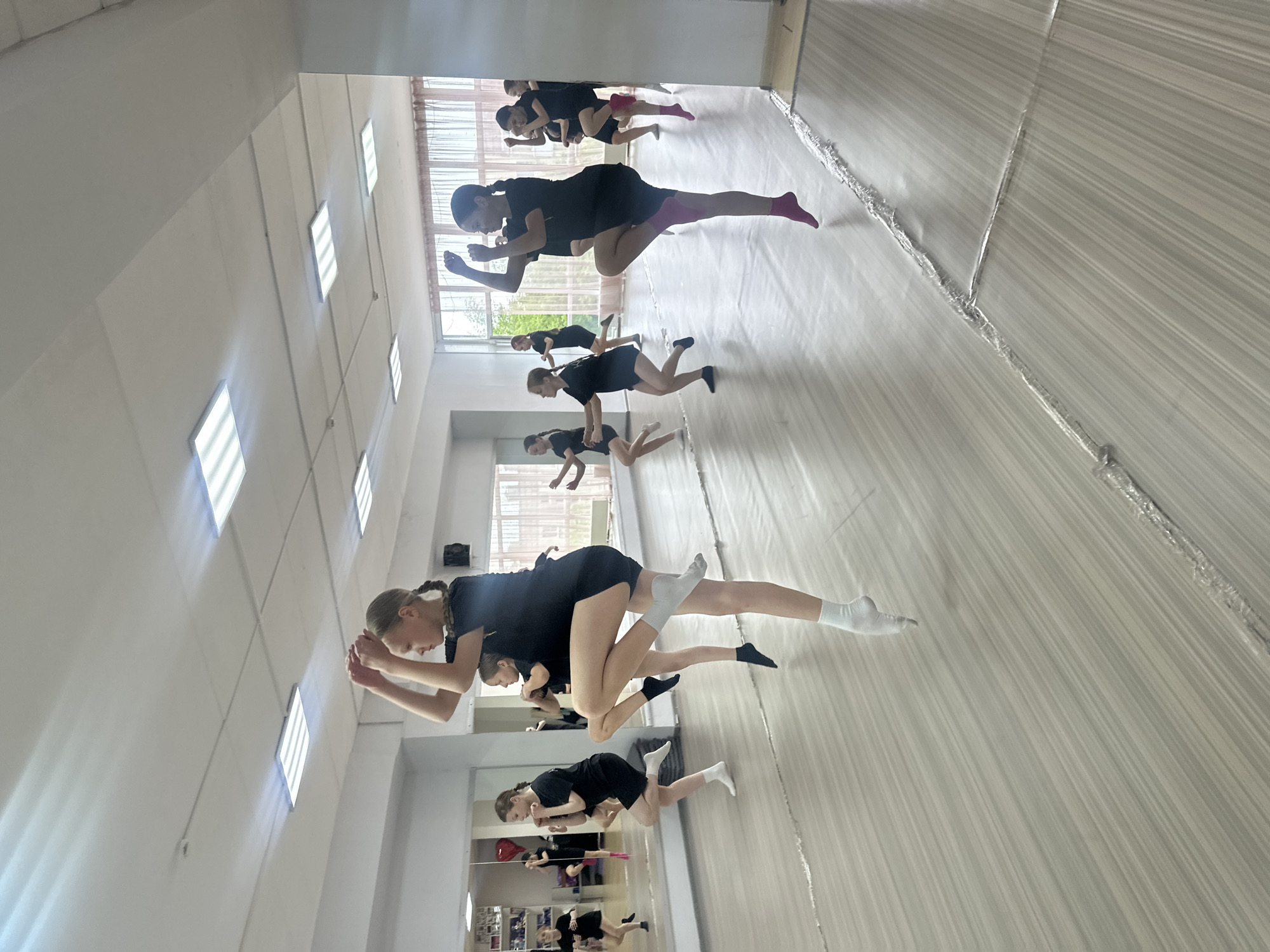The image shows a group of young women in a dance studio, practicing what appears to be a choreographed routine. They are all wearing black leotards and shorts, with some wearing different colored socks. The room has a light-colored wooden floor and a mirrored wall. There are large windows with blinds, and fluorescent lights on the ceiling. The overall atmosphere seems focused and energetic, with the dancers engaged in synchronized movements. The pose of the two main subjects suggests a lift or a dynamic partnering element within their dance. The time of day is likely daytime due to the natural light coming from the windows. No visible text is present in the image. The location is Votkinsk, Russia.