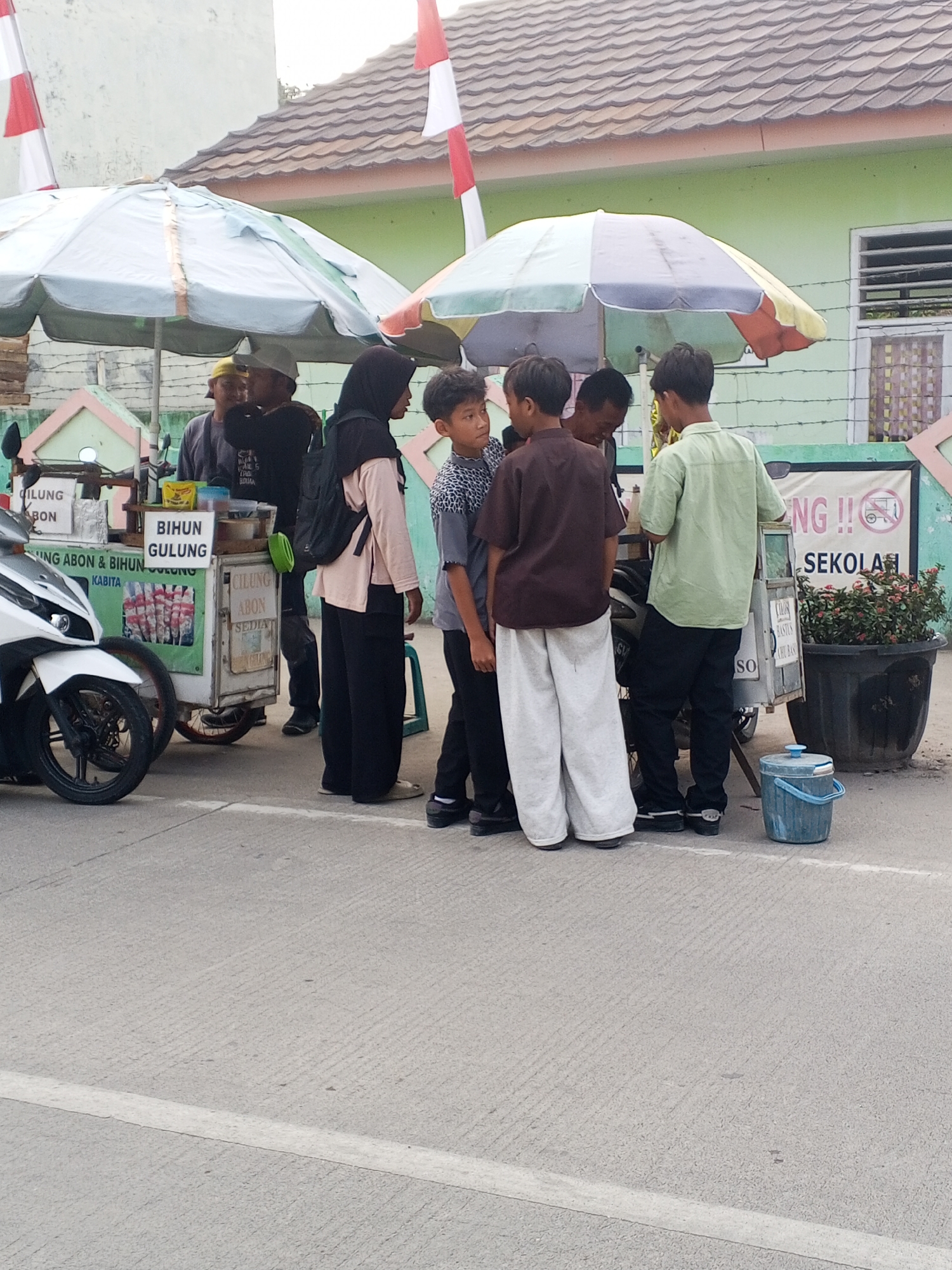 In the city of Ciruas, Indonesia, a street scene unfolds with a food cart selling "Bihun Gulung" and "Cilung Abom" under a colorful umbrella. A white motorcycle is parked to the left of the cart. Several individuals are gathered around the cart, seemingly engaged in conversation or making a purchase. The background shows a light green building with a tiled roof and a fence. Two Indonesian flags, red and white, are visible on poles near the building. The ground is paved asphalt. The time of day appears to be daytime, possibly late afternoon due to the lighting.