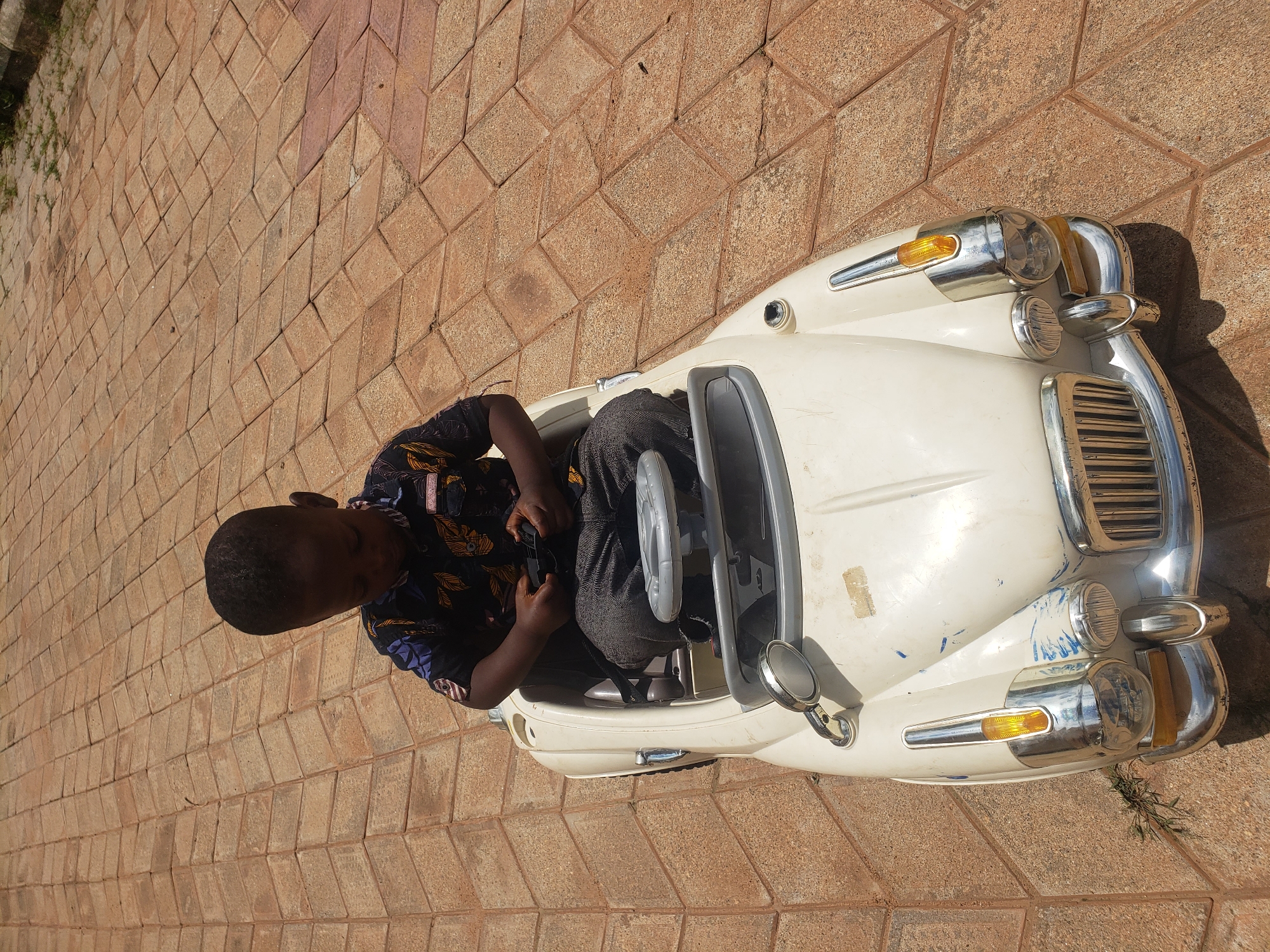A young child with dark skin and short dark hair sits in a white toy car on a paved ground. The child is wearing a dark shirt with a colorful floral pattern and dark pants. The child is holding a black object in their hands, possibly a toy steering wheel or remote control. The toy car is detailed with chrome accents, including a grille, headlights, and side mirrors. The ground is paved with interlocking brown and tan bricks arranged in a herringbone pattern. In the background, a dark border runs along the edge of the paved area, with some green vegetation visible beyond it. The scene appears to be outdoors in a bright, sunny environment. The location is identified as Sabon Tasha, Nigeria.