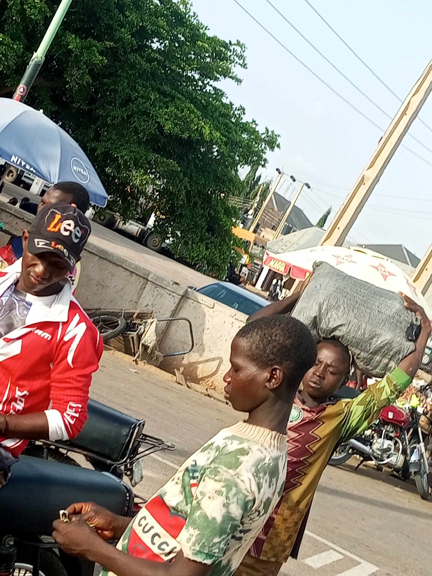 A group of people, including at least two young boys and a man, are gathered outdoors, likely in a market or busy street. The man in the foreground wears a red and white jacket and a black cap with the word "Lee" visible on it. He is on a motorcycle. To his right, two young boys are visible. One, in the center of the frame, wears a camouflage-patterned shirt and holds a large bundle on his head, supported by an umbrella. The other boy is closer to the viewer and wears a shirt with a red heart design and the word "CUCCI". In the background, there is an umbrella with "NIVEA" printed on it, some buildings, trees, and a clear sky, suggesting it is daytime with fair weather. There are also motorcycles and other vehicles visible, indicating a potentially bustling environment. The overall scene appears to be one of daily commerce or transport in what could be a town or city.