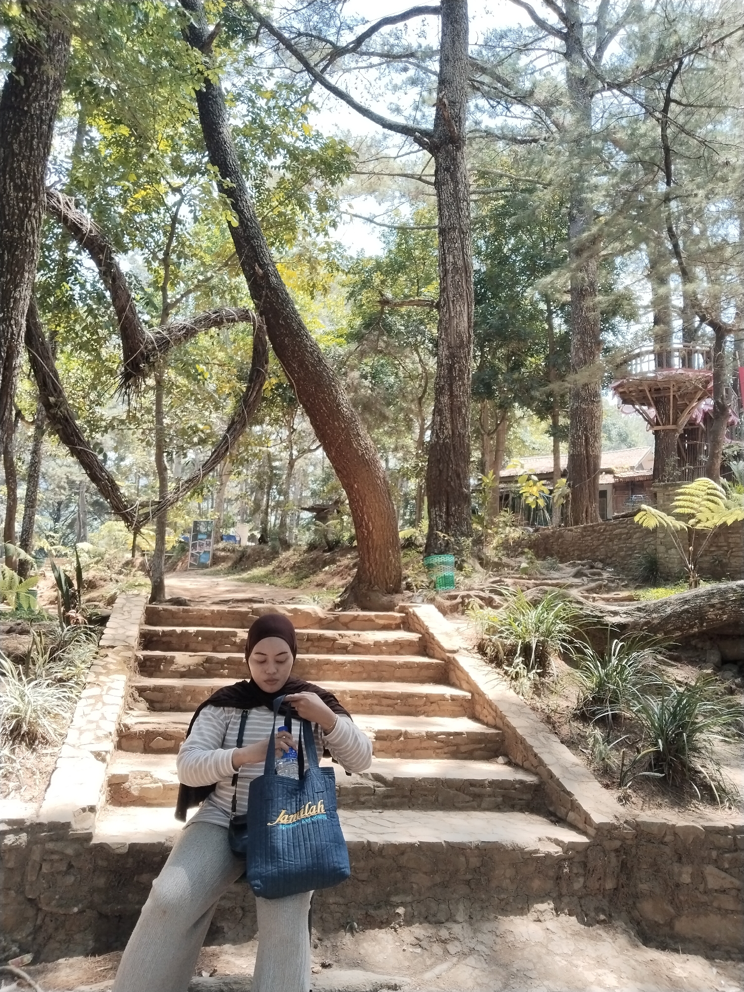 A person wearing a hijab and a striped shirt sits on a stone staircase in a park setting. The person is holding a dark blue tote bag with "Jamilah" embroidered on it in yellow, and a bottle of water. The staircase is surrounded by trees and vegetation, with a wooden structure visible in the background. The location appears to be Kuwukan, Indonesia, as suggested by the prompt.
