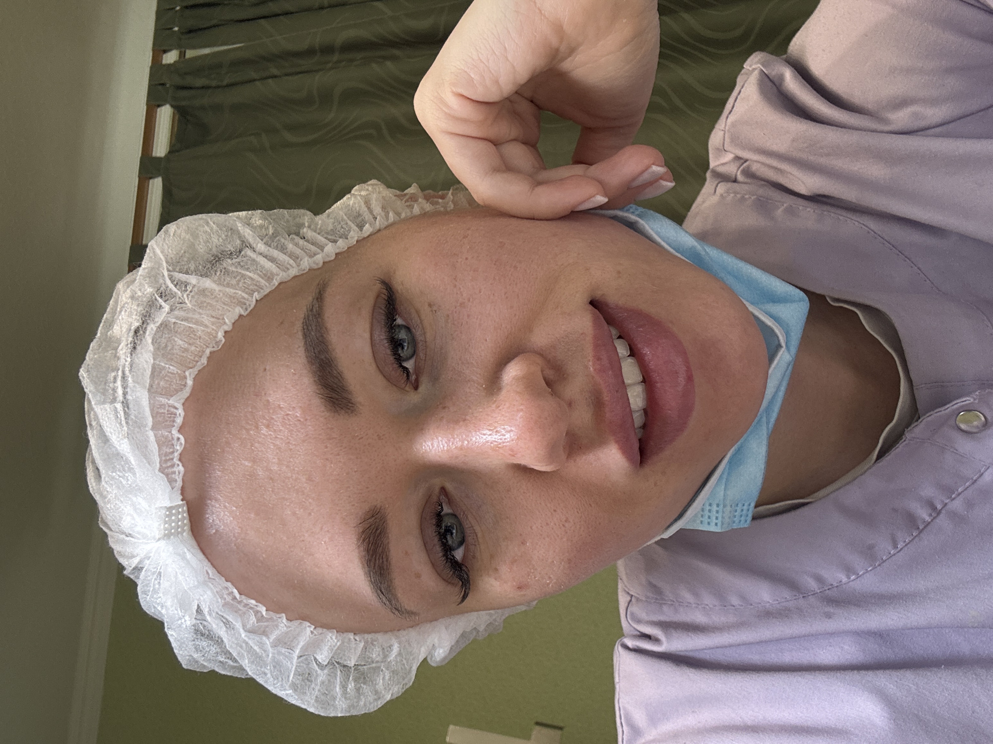 A person wearing a white disposable cap and a light purple scrub top is visible in a close-up, angled shot. A light blue surgical mask is pulled down to the side, revealing the person's face. The person is touching their cheek with their fingers. The background includes a green wall with window blinds. The image was taken in Ферма, Russia.