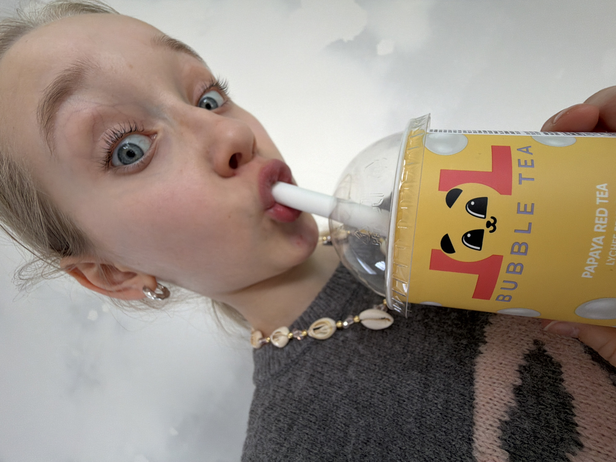 A fair-skinned girl with blue eyes is drinking bubble tea. She is wearing a gray sweater with pink stripes and a shell necklace. She has a silver hoop earring in her left ear. The bubble tea cup is yellow with a cartoon face and the words "L-L Bubble Tea" and "Papaya Red Tea". The background is a blurry white and gray pattern. No location information is discernible from the image.