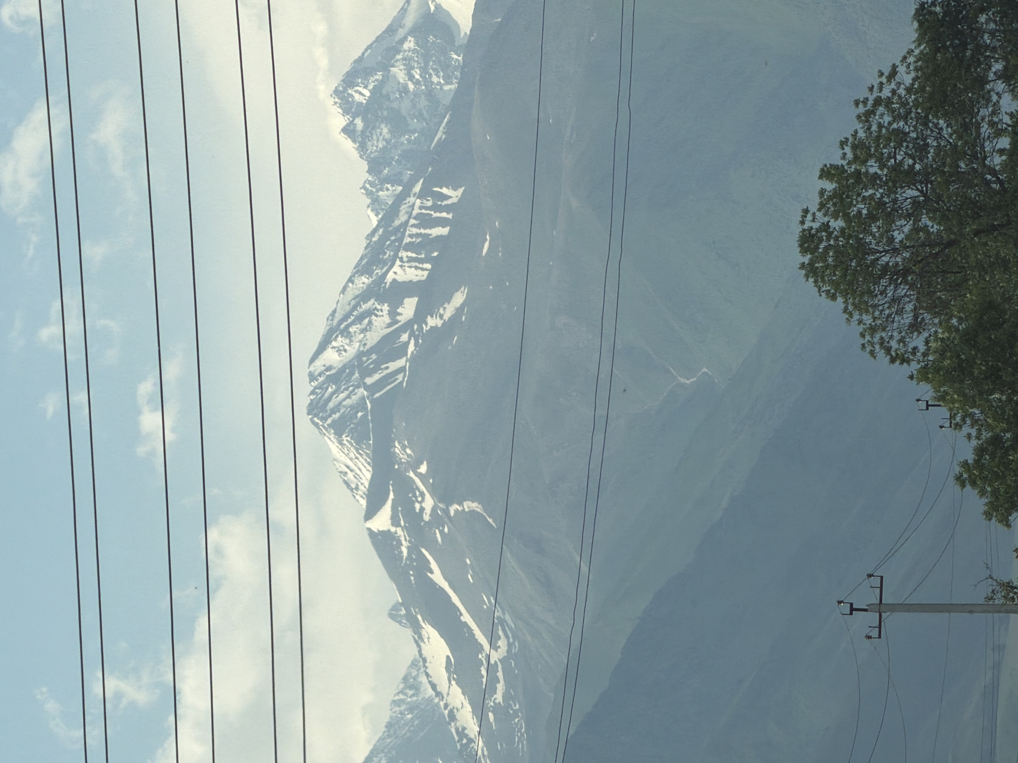 The image shows a wide shot of a mountain range under a partly cloudy sky, with power lines running horizontally across the frame. The mountains in the background are snow-capped and appear rugged. In the foreground, there are trees and utility poles. The scene is likely outdoors, possibly a rural or mountainous area, given the presence of power lines and the natural landscape. The lighting suggests it is daytime.