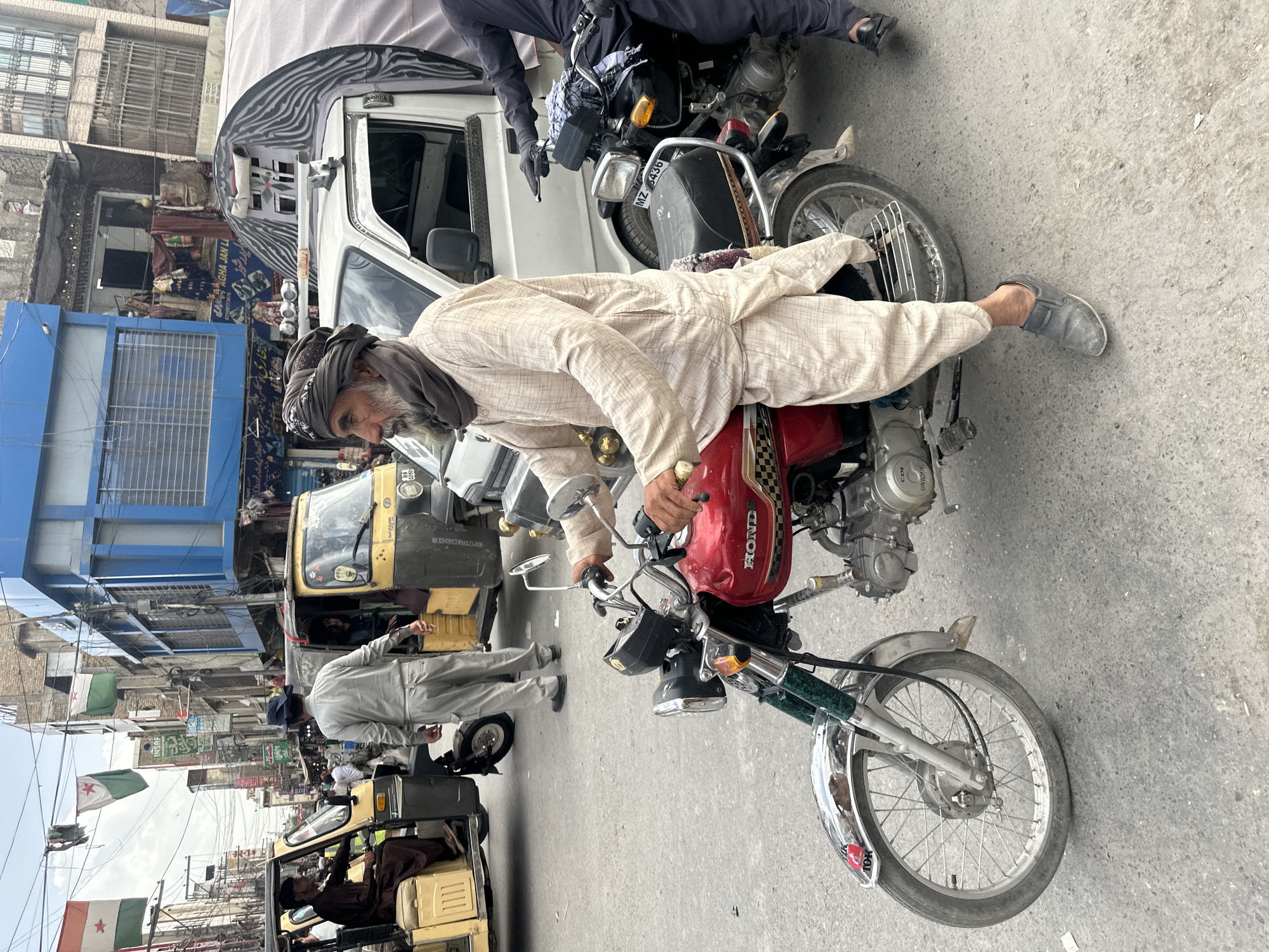 The image depicts a street scene in Quetta City Tehsil, Pakistan, featuring an elderly man in traditional clothing lying across the seat of a red Honda motorcycle. He is wearing a turban and light-colored clothing. The motorcycle is parked on a paved street alongside other vehicles, including auto-rickshaws and a white van. The background includes buildings with signs written in Urdu. Political flags, including those with crescent and star symbols, are visible in the distance, attached to buildings or utility poles.