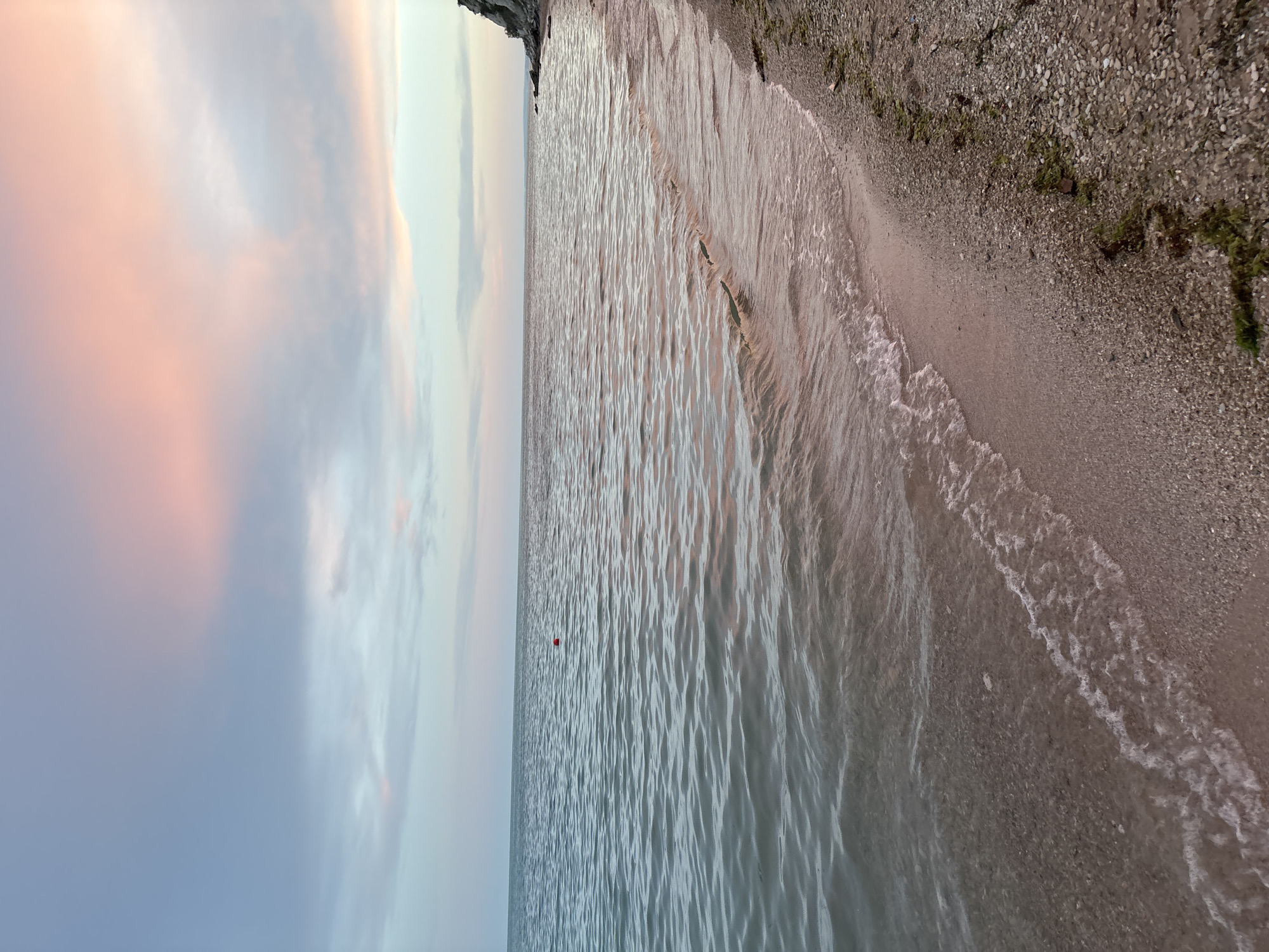The image depicts a serene beach scene at dusk in Topola, Bulgaria. The sky is painted with soft hues of pink and blue from the setting sun, with wispy clouds stretching across the horizon. The calm sea reflects the muted colors of the sky, its surface rippled by gentle waves lapping onto the shore. The beach itself is composed of fine sand and pebbles, with some scattered green seaweed along the tideline. In the distance, a rocky cliff formation marks the edge of the coastline. A small, red buoy is visible in the water. The overall atmosphere is tranquil and peaceful, capturing the quiet beauty of the Black Sea coast.