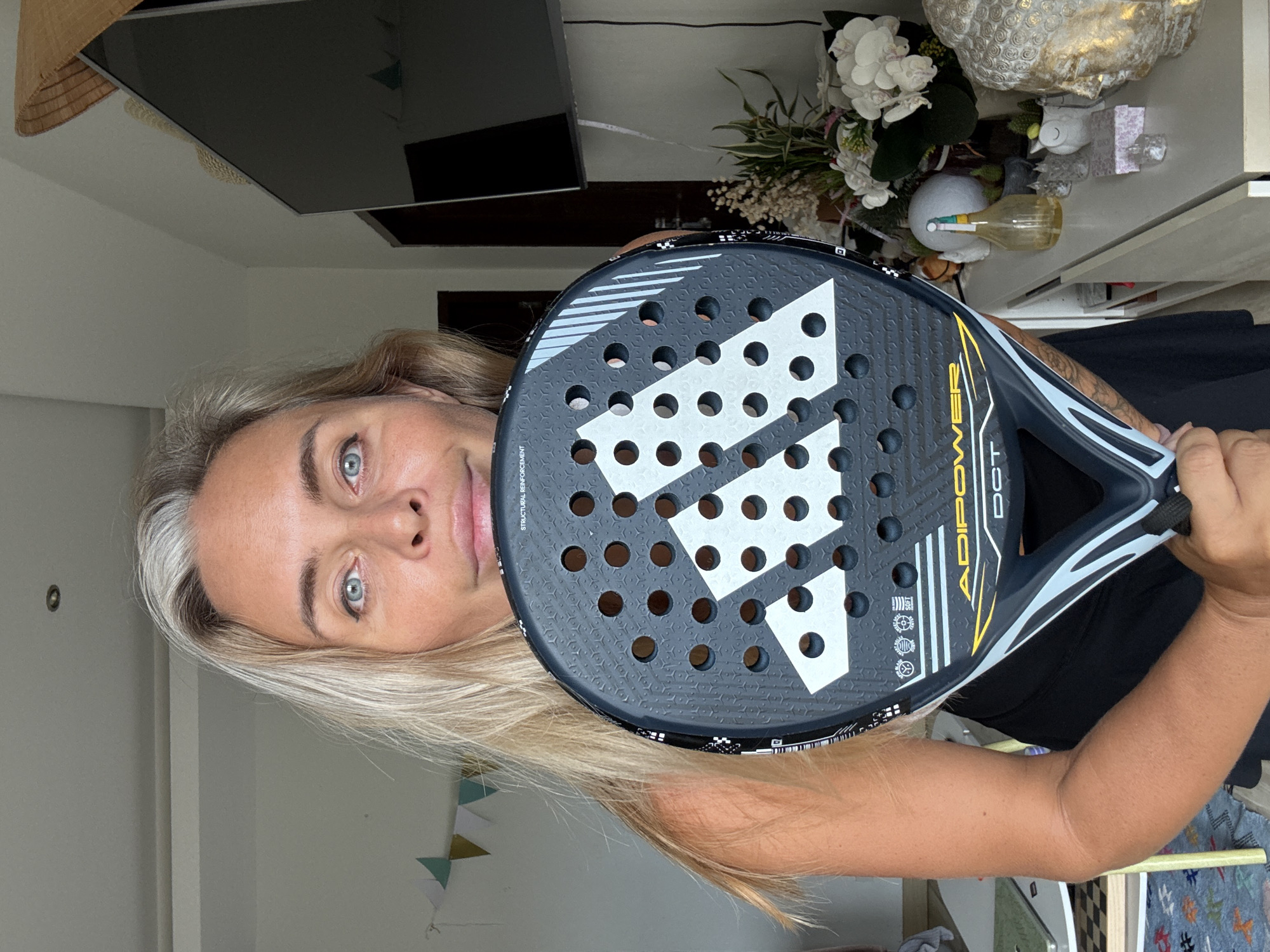 A medium shot shows a blonde woman with blue eyes holding an Adidas padel racket in front of her face. She is wearing a black tank top. The background is an indoor setting with a TV, a shelf with decorative items, and a wall with hanging triangular flags. The lighting suggests it is daytime. The location is Ko Samui District, Thailand.