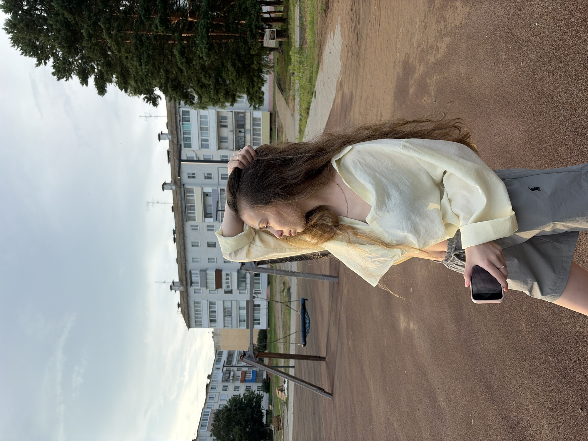 A young woman with long, brown hair stands in an outdoor playground in Noviy, Russia. She is wearing a light yellow oversized shirt and gray shorts. Her right hand is raised to her head, and she is holding a smartphone in her left hand. The playground has a brown, sandy surface and a wooden swing set with a blue seat. In the background, there are several multi-story apartment buildings and a large evergreen tree. The sky is overcast.