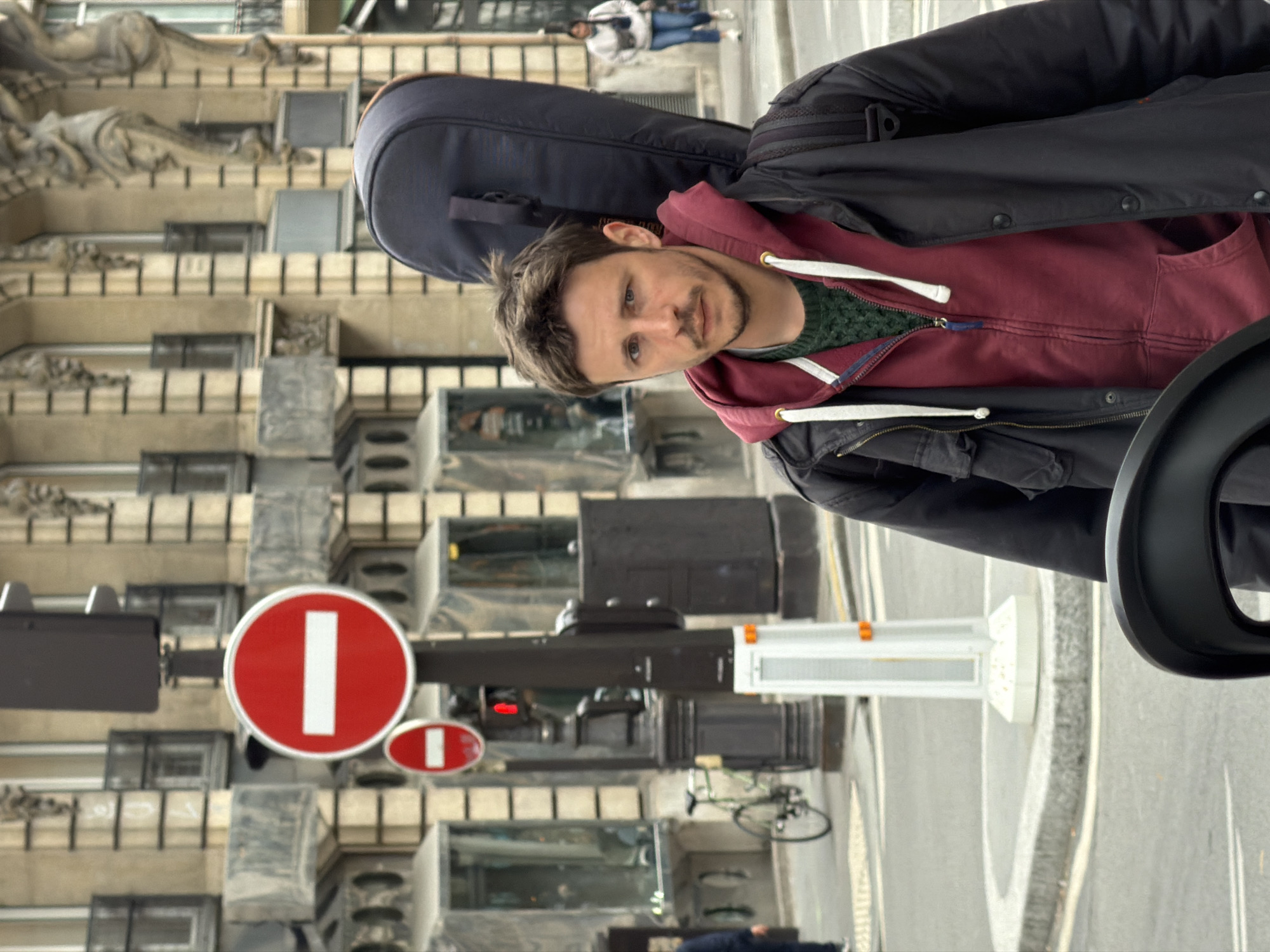 A medium, eye-level, rotated shot depicts a man in Paris, France. He has a dark blue musical instrument case on his back. He is wearing a black jacket and maroon hoodie. Behind him is a building with ornate architectural details. A red-and-white "no entry" traffic sign is visible. A bicycle is parked in the background. Pedestrians walk in the distance.