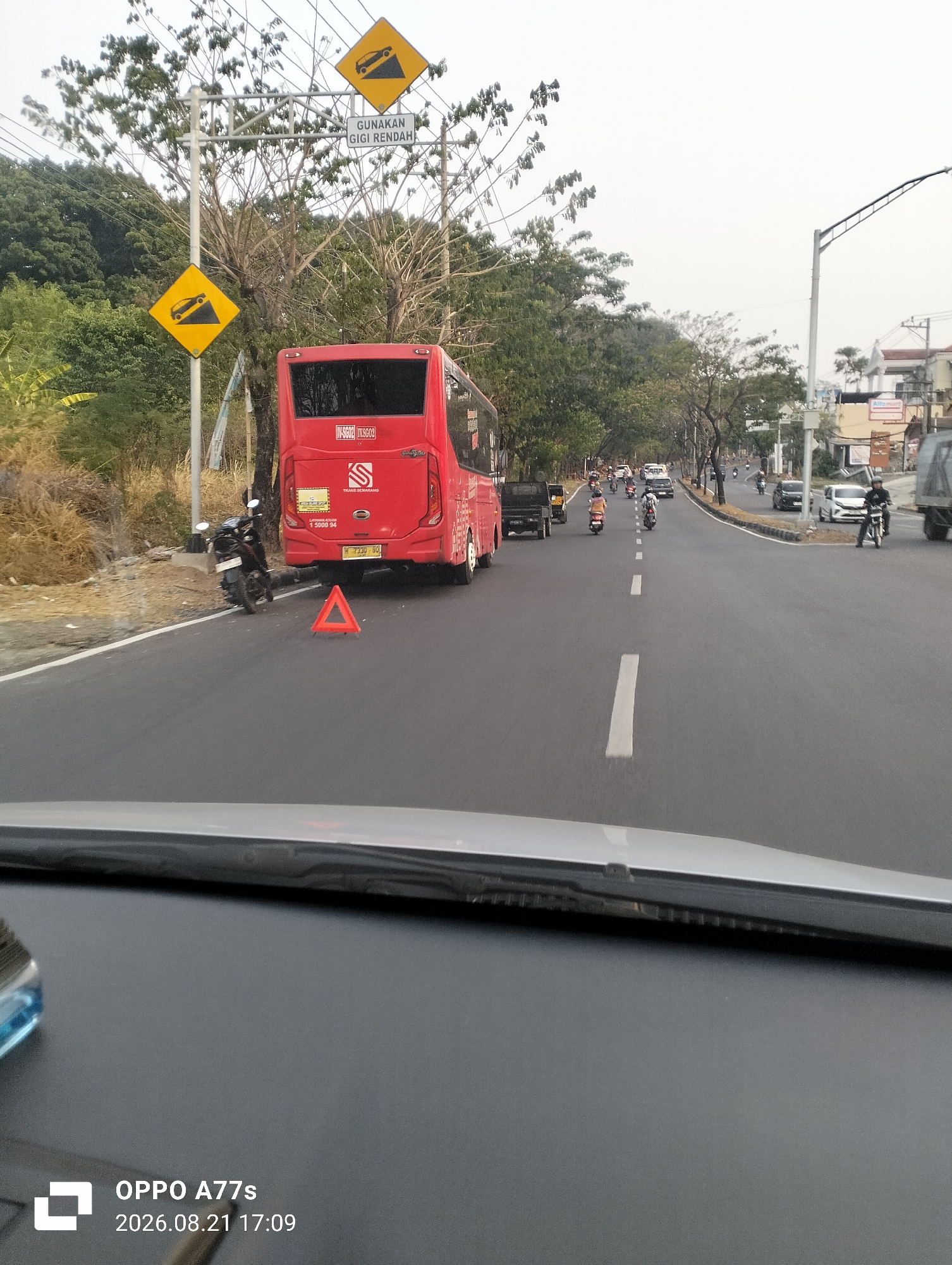 The image depicts a street scene in Semarang, Indonesia. A large red bus with the text "TRANS SEMARANG" is parked on the right side of the road. A motorcycle is parked behind it. An orange triangular warning sign is placed on the road in front of the bus. Several other vehicles, including cars and motorcycles, are visible on the road, suggesting active traffic. Traffic signs indicating a steep incline and a requirement to use a low gear are mounted on poles in the background. The environment appears to be a road in an urban or suburban area, with trees and buildings visible along the sides. The sky is overcast.
