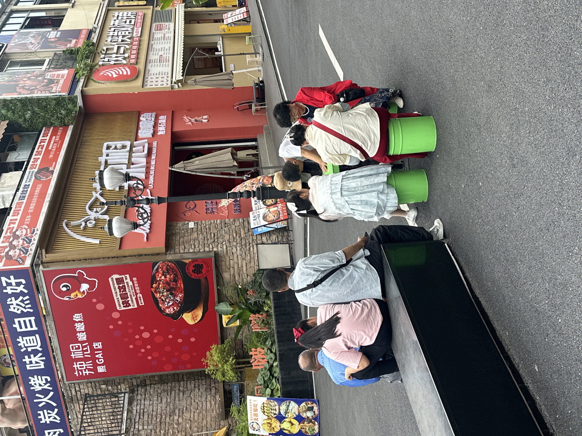The image shows a group of people seated on green buckets and a bench on a city street in Chongqing High-tech Zone, China. The scene includes an elderly woman in a red shirt, a child with a red ribbon in their hair, and other adults in casual clothing. They appear to be engaged in a conversation or activity together. The background features storefronts with Chinese signage and advertisements for restaurants, including "La Xiang Bo Bo" and "Xi Gai Store". A large red poster advertises "Spicy and Delicious Fish." The street is paved with gray asphalt. The lighting is overcast.