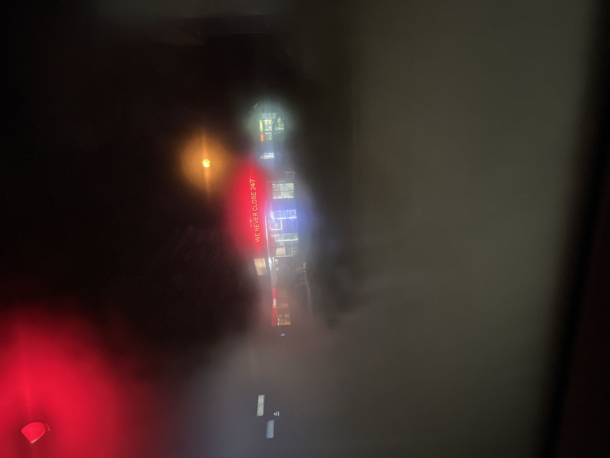 A dark, hazy, horizontally-oriented night shot, taken at low resolution. A bright orange light and a blurry red light are visible. The illuminated red letters spelling "WE NEVER CLOSE 24/7" are oriented vertically. In the top right, blurred windows can be made out. A street appears in the lower portion of the image, with a few rectangular light reflections along its length. The location may be in Plovdiv, Bulgaria.