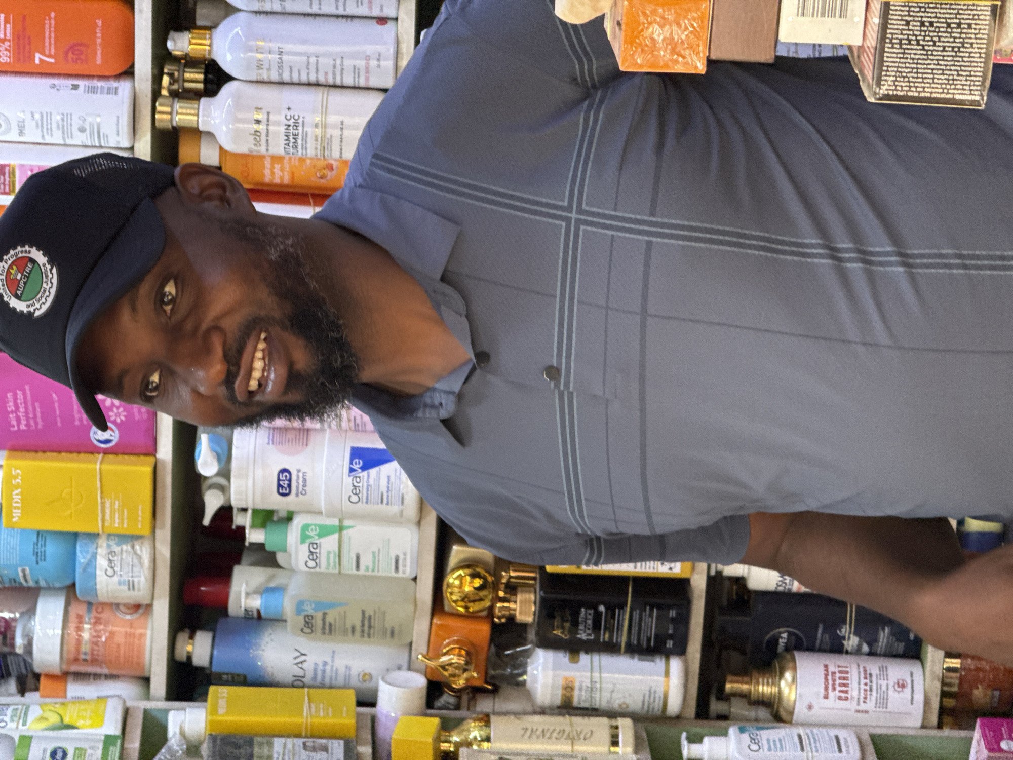 The image depicts a man with a black beard, wearing a black baseball cap with an emblem, standing in front of shelves stocked with numerous skincare and cosmetic products. The man is smiling and appears to be looking directly at the camera. The products on the shelves include various brands such as Cerave, Olay, and others with labels like "Vitamin C + Turmeric," "White Carrot," and "Licorice." The text "United for Progress" and "AUPCTRE and Social Justice" is visible on the emblem of the baseball cap. Based on the products and appearance of the environment, this could be a retail setting selling beauty and skincare items, potentially in Abuja, Nigeria.