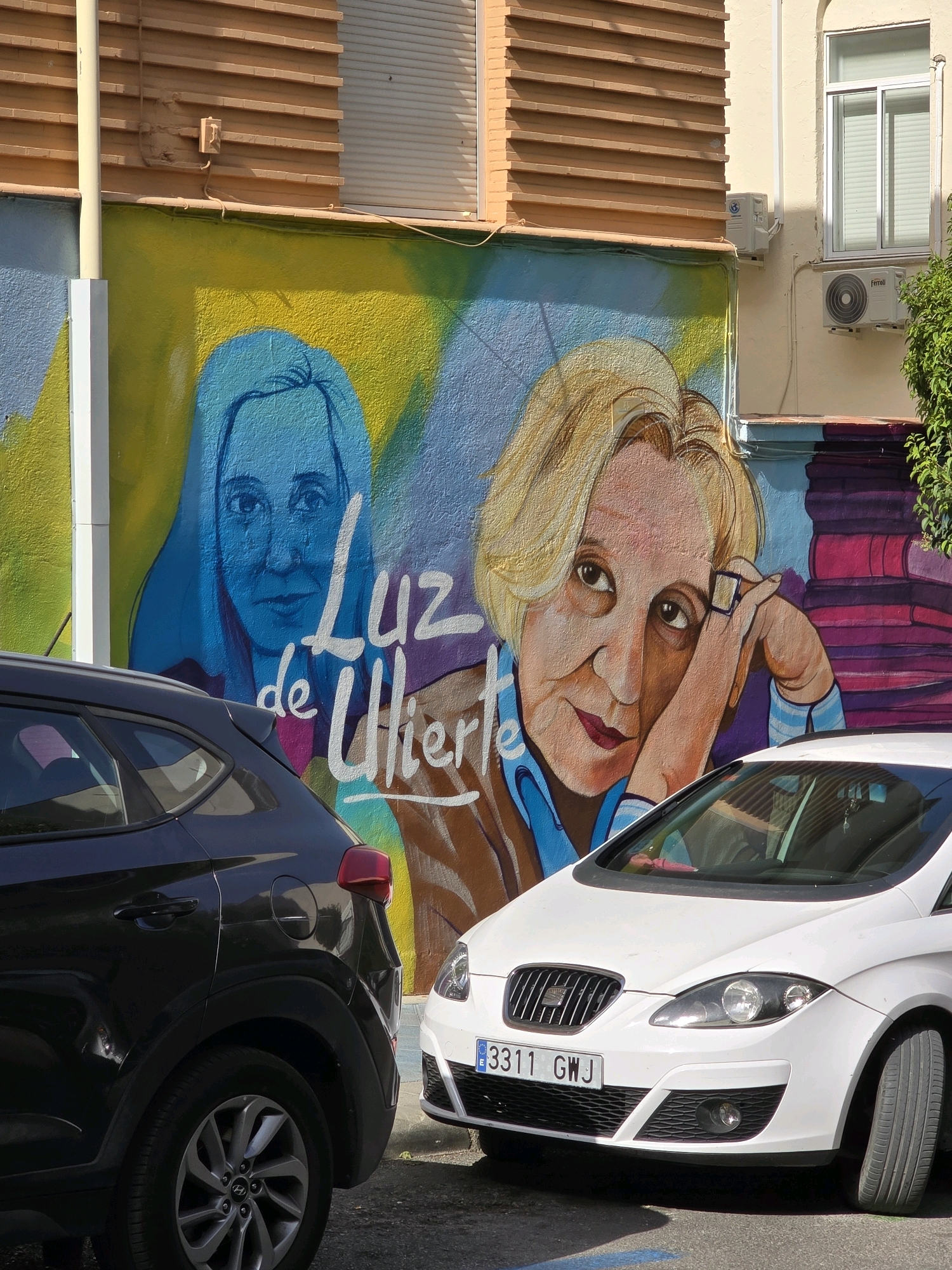 This image depicts a mural on a building in Jaén, Spain, featuring two portraits of women. The mural is vibrant, with the left portrait rendered in blue outlines against a yellow and blue background, and the right portrait in warmer tones of orange and yellow. White lettering, partially obscured, reads "LUZ de ULIERTE". The mural is situated on a building with a textured, tan upper facade and cream-colored walls below, featuring air conditioning units and a window. In the foreground, two cars are parked: a dark-colored SUV on the left and a white hatchback on the right. The white car's license plate reads "3311 GWJ". The scene appears to be an outdoor urban setting, with a shadow cast across the foreground.