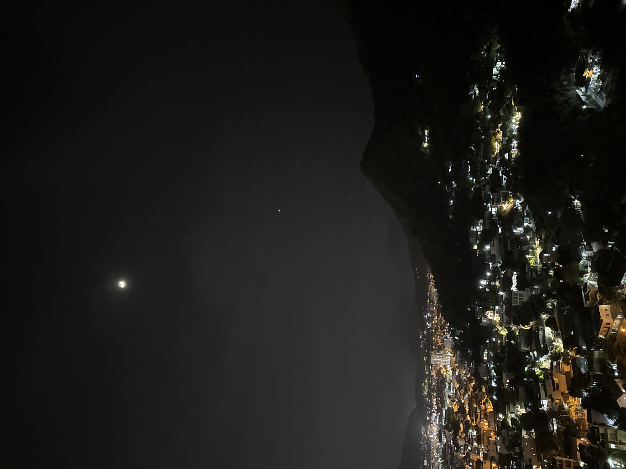 The image is a nighttime aerial view of a densely populated hillside in Bac Nha Trang Ward, Vietnam. The scene is illuminated by the lights of numerous houses and buildings that cling to the steep slope. The lights are a mix of warm yellow and bright white, creating a twinkling effect against the dark landscape. Trees are interspersed among the structures, their foliage appearing dark silhouettes against the artificial lights.
In the upper left portion of the image, the night sky is visible. A bright, luminous object, likely the moon, is prominent, casting a soft glow. Another smaller, brighter point of light, possibly a planet or a star, is also visible nearby. The sky is otherwise dark, suggesting a clear night.
The overall impression is one of a vibrant community nestled within a natural, mountainous setting. The perspective suggests the photo was taken from a high vantage point, possibly from a drone or a helicopter, looking down and across the hillside. There is no visible text in the image. The time of day is clearly night, and the clear sky suggests fair weather. The location cues, like the dense hillside settlement, are characteristic of many coastal towns in Vietnam.