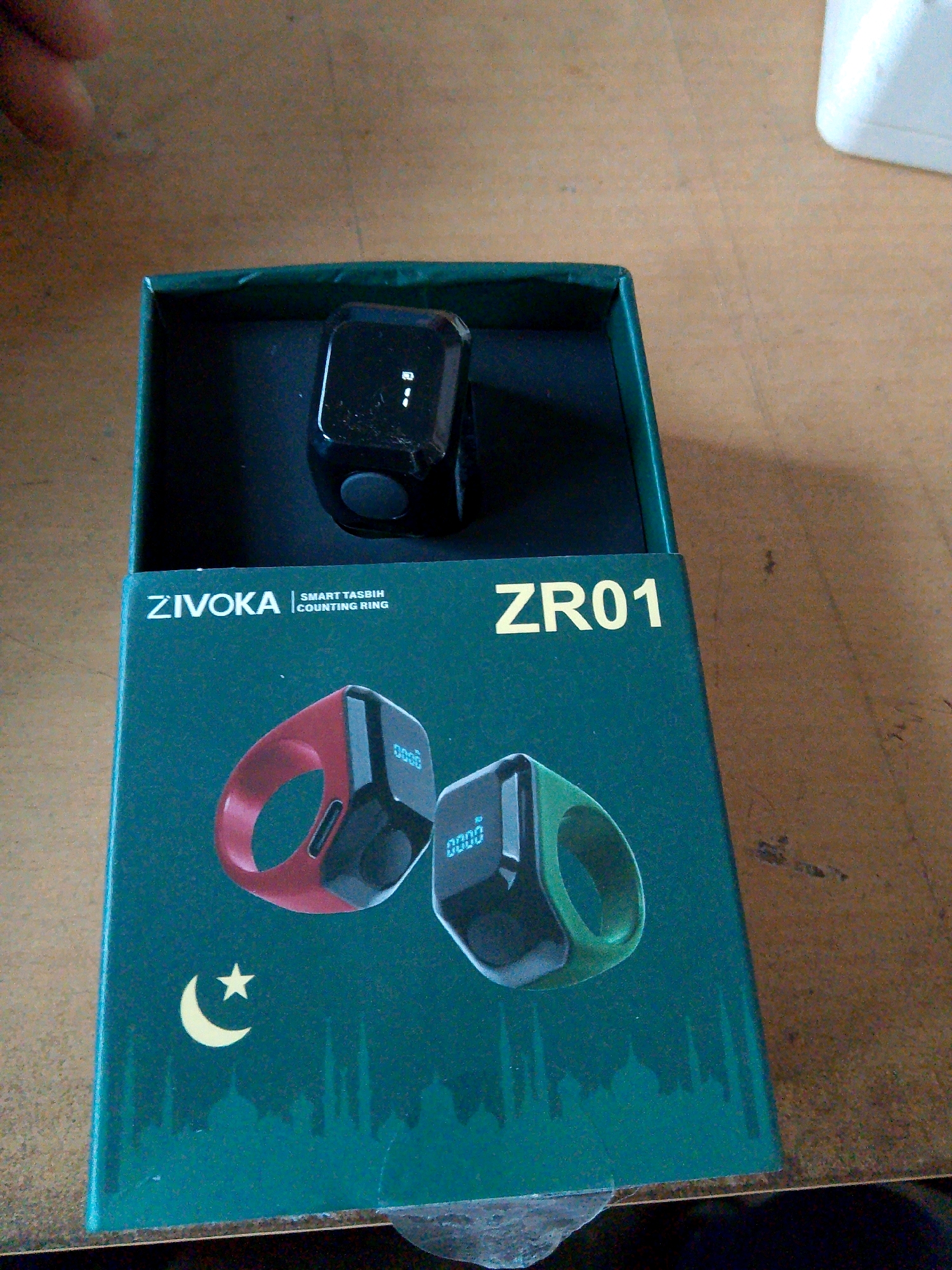 In an indoor setting, on a table, is a dark green product box for a "ZIVOKA SMART TASBIH COUNTING RING" with the model number "ZR01." Inside the box, a black digital tasbih ring with a small screen is visible. The box also features images of a red and a green tasbih ring, a crescent moon and star, and silhouettes of mosque domes. The image was taken in Kuripan Utara, Indonesia.