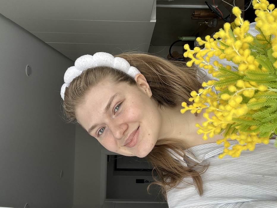 A young Caucasian female subject is depicted from the chest up, positioned indoors in a residential setting in Nikol'skii, Russia. Her light-colored hair is pulled back, secured by a white, puffy, cloud-shaped headband. She looks directly towards the viewer with a subtle smile, exhibiting light-colored eyes. She is wearing a light-colored, possibly striped, collared shirt. In the immediate foreground, she holds a bouquet of artificial bright yellow flowers resembling mimosa, characterized by numerous small yellow florets and green fern-like leaves. The background on the left features a partial doorway leading into a darkened room. On the right, a segment of a modern kitchen or living area is visible, including white overhead cabinetry and a dark countertop. Objects present on the countertop consist of a green bottle, a metallic gold-colored decorative item, a clear glass bottle, and several black electronic cables. The ceiling is a light gray, fitted with two visible recessed lighting fixtures.