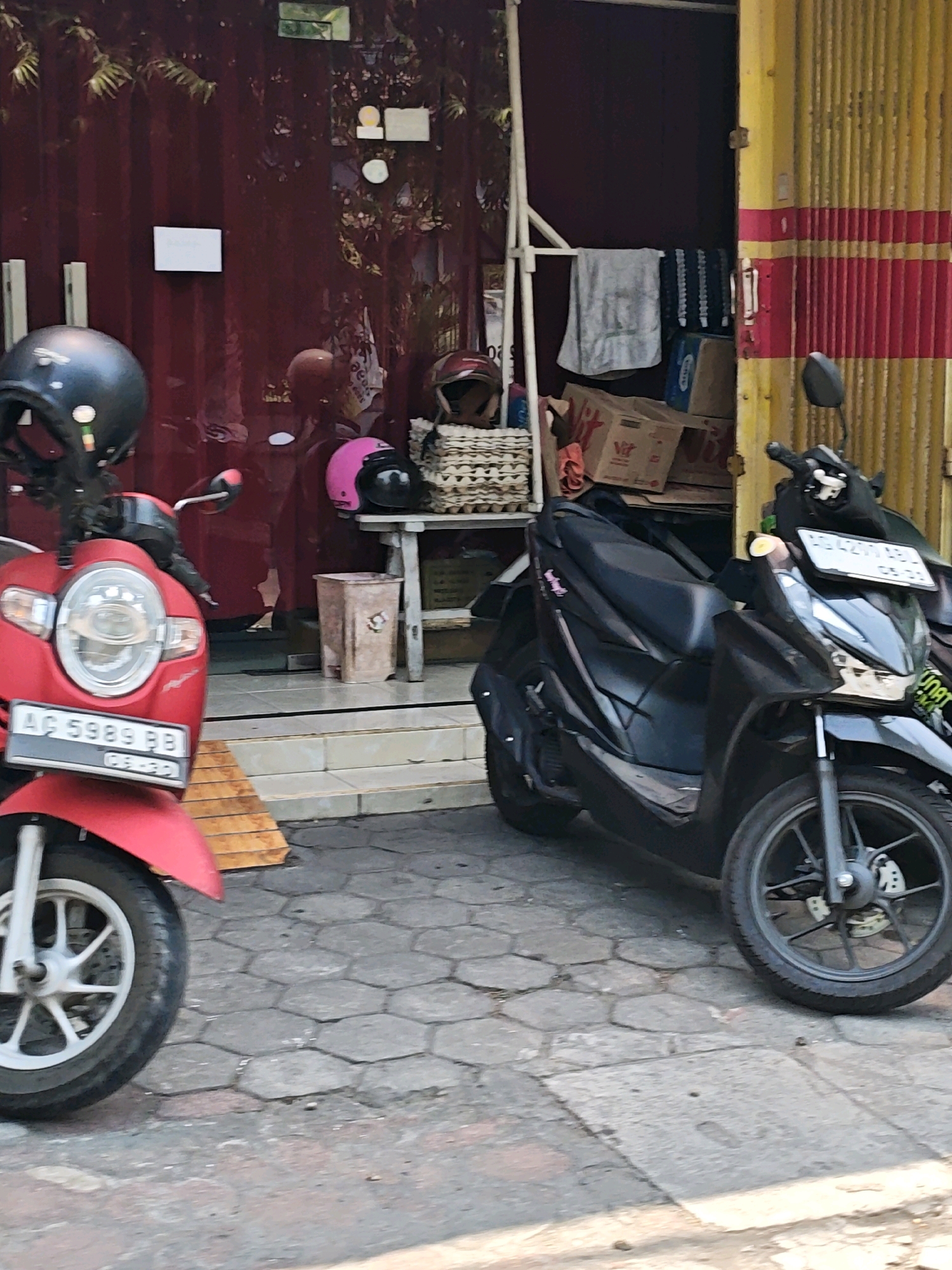 Two scooters are parked on a paved street in front of a building with a red corrugated metal wall and a yellow and red striped rolling door. The scooter on the left is red with a black helmet on its handlebars and a license plate reading "AG 5989 BB". The scooter on the right is black with a white license plate. There are several boxes and a woven basket on a wooden stand behind the scooters, and a pink helmet is visible on the stand. A trash can is also present near the entrance of the building. The ground is paved with hexagonal stones. The scene appears to be in Kediri City, Indonesia, indicated by the license plate format.