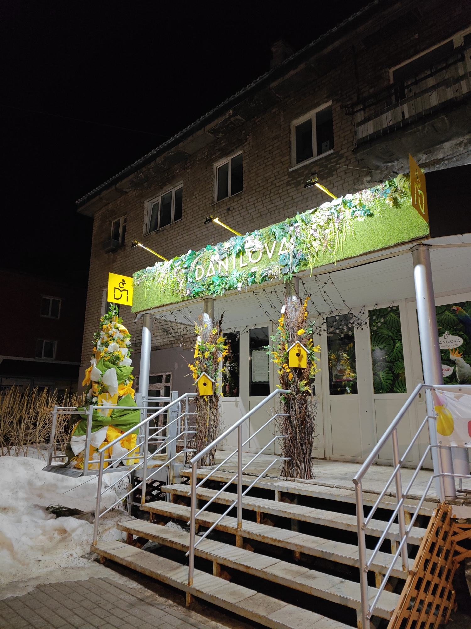 The image captures the exterior of the "Danilova" establishment in Chaikovsky, Russia, at night. The building is a two-story brick structure with lit windows on the upper floors. The entrance is elevated and accessed by a set of wooden stairs with silver metal railings.
The entrance is decorated with green and floral arrangements, including a sign with the name "Danilova." Two poles flank the entrance, adorned with decorative birdhouses and twigs. To the left of the stairs, there's a decorative arrangement with yellow, white, and green bows.
The ground is covered with snow. The scene is illuminated by artificial lights, including those under the awning above the entrance.