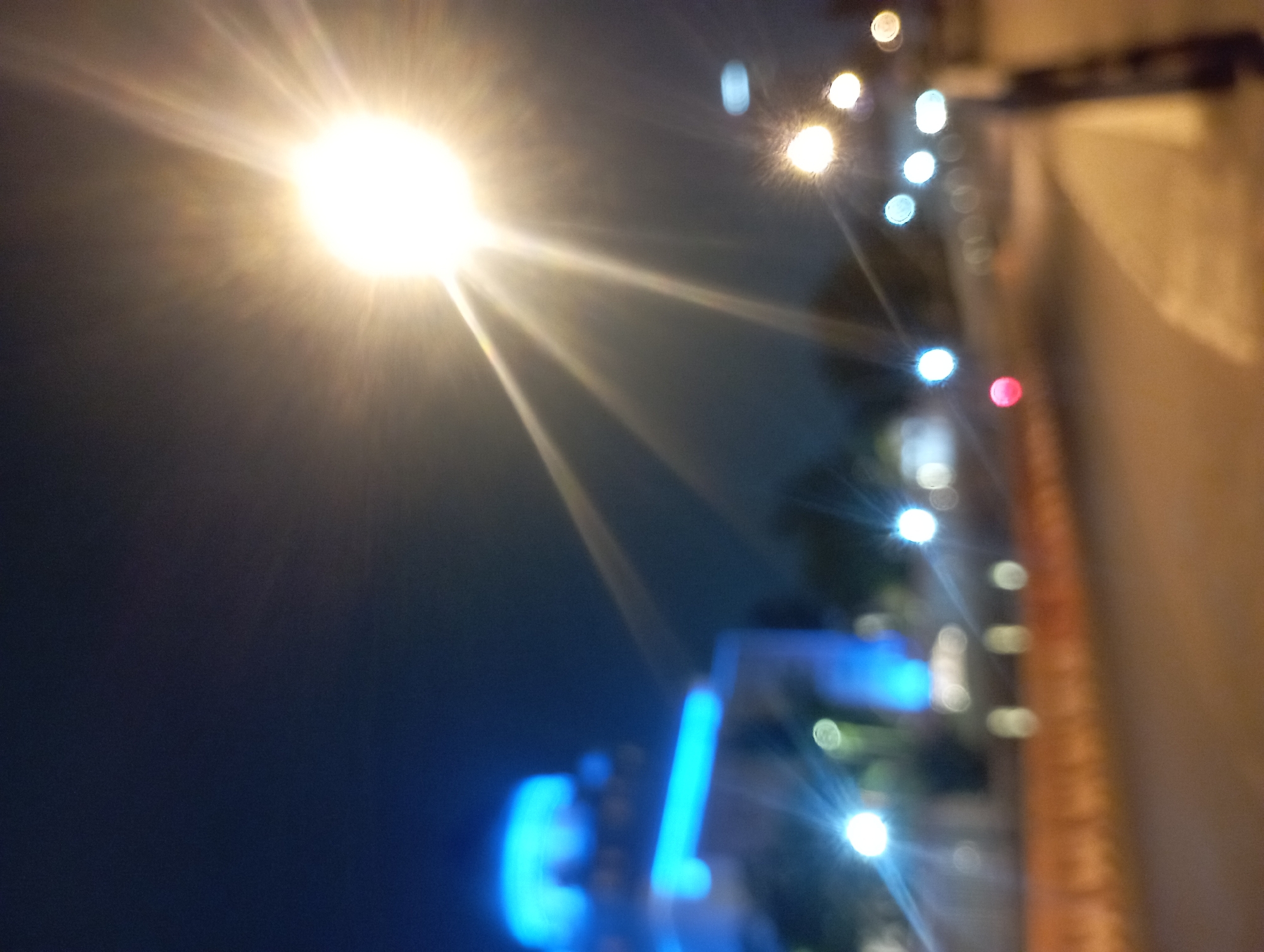 The image is a night-time, out-of-focus photograph taken on a street in Surakarta, Indonesia. A bright street lamp with prominent light rays dominates the upper right portion of the frame. In the background, a building is illuminated with blue lights, possibly a mosque or other public structure. The street itself is visible in the lower portion of the image, with several blurred lights suggesting other vehicles or streetlights. The overall impression is a bustling urban environment at night.