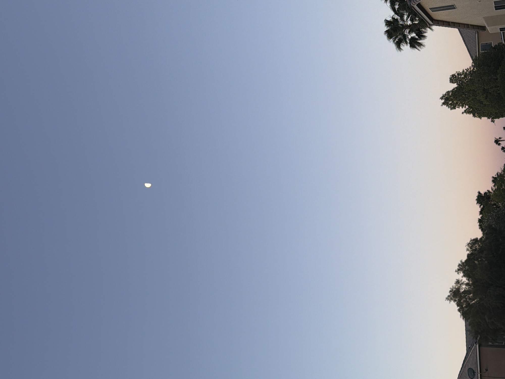The image shows a sky gradient from blue to orange, suggesting either sunrise or sunset. A crescent moon is visible in the blue portion. Trees and a building are visible on the right edge. The location is possibly Las Vegas, United States of America, although no definitive evidence is present in the image. No people, subjects, actions, or interactions are depicted.