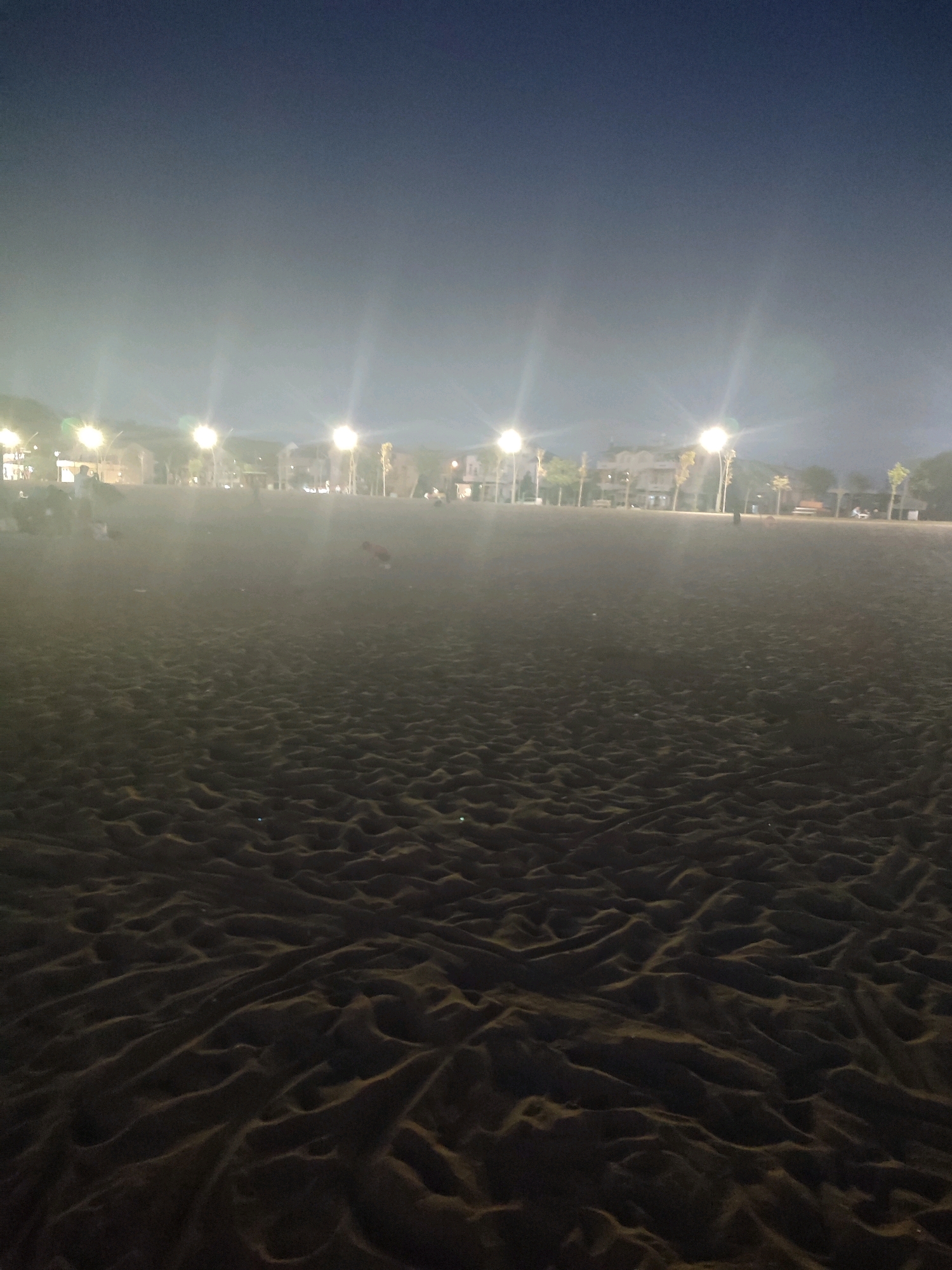 The image depicts a beach at night in Kandıra, Turkey, illuminated by streetlights and ambient light. The foreground is dominated by the textured surface of the dark sand, showing ripples and undulations. In the mid-ground, a row of streetlights casts a glow across the beach, illuminating a promenade or walkway bordering the sand. Buildings and trees are visible in the background, suggesting an urban or semi-urban coastal setting. A few indistinct figures can be seen scattered along the promenade, some appearing to be sitting or walking. The sky above is dark and clear, transitioning to a lighter hue towards the horizon where the light sources are located. The overall atmosphere is calm and serene, typical of a beach at night.