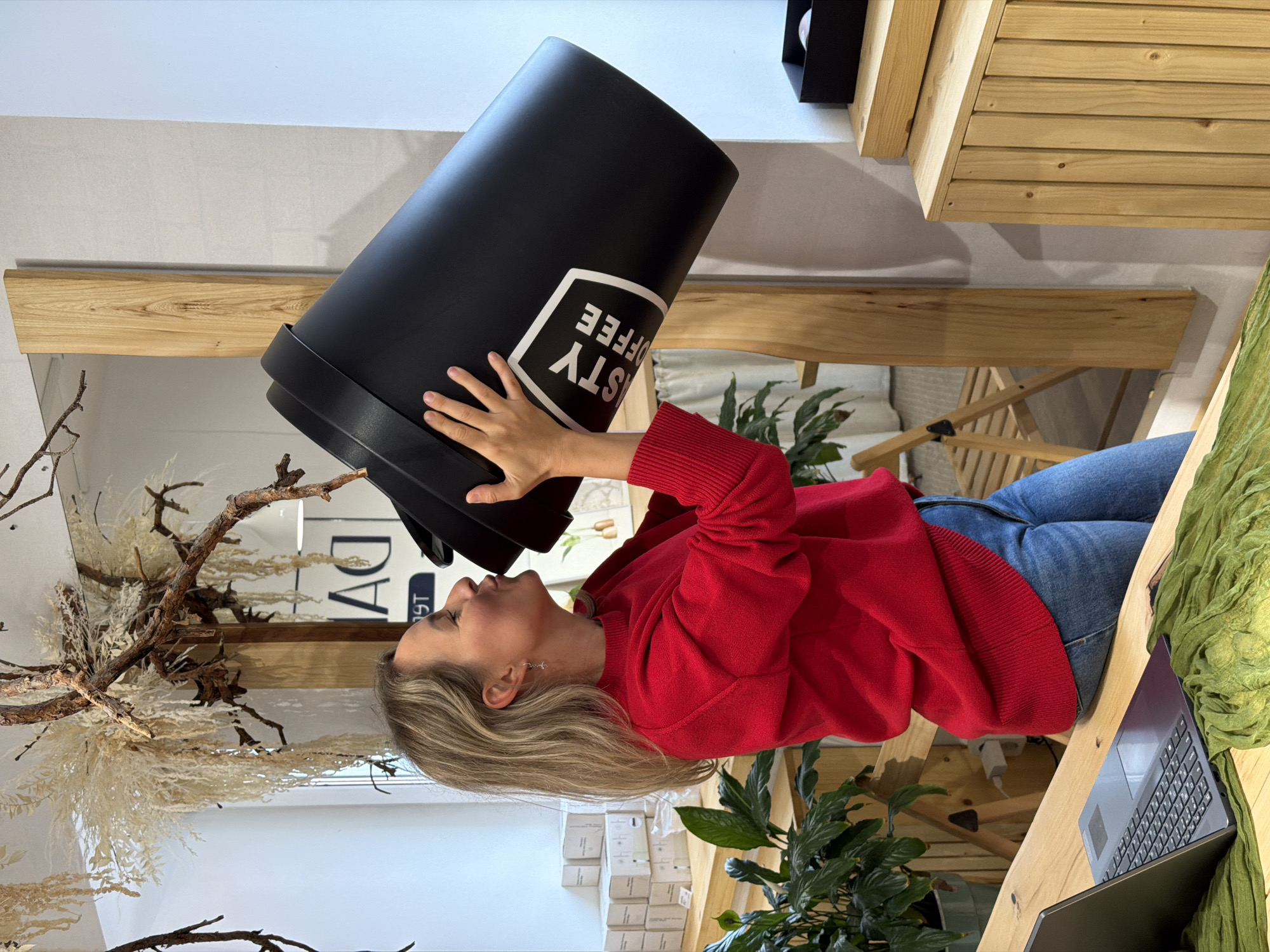 A woman in a red sweater and blue jeans holds an oversized black coffee cup to her mouth. The cup has a white logo. She's indoors, near a wooden table with a laptop and a green scarf on it. There are plants, wooden shelves, and decorative branches in the background. The boxes stacked behind her indicate that this could be a store. The location is Chaikovsky, Russia.