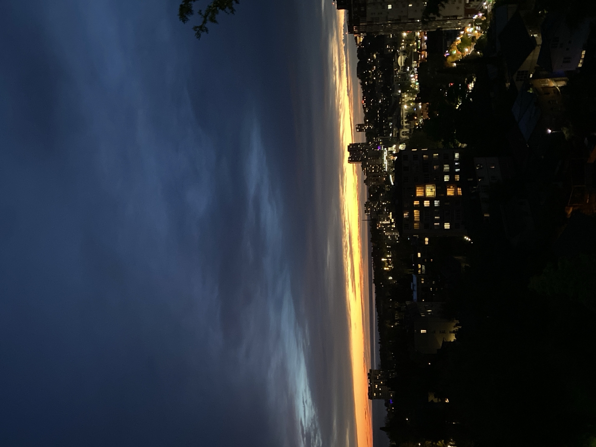 The image captures a twilight scene in Sochi, Russia. On the left side is the evening sky with shades of blue transitioning into a vibrant orange horizon. The right side reveals a cityscape dotted with lit buildings. Dark areas suggest dense foliage or unlit structures closer to the foreground. The scene appears to be shot from a higher vantage point, overlooking the city.