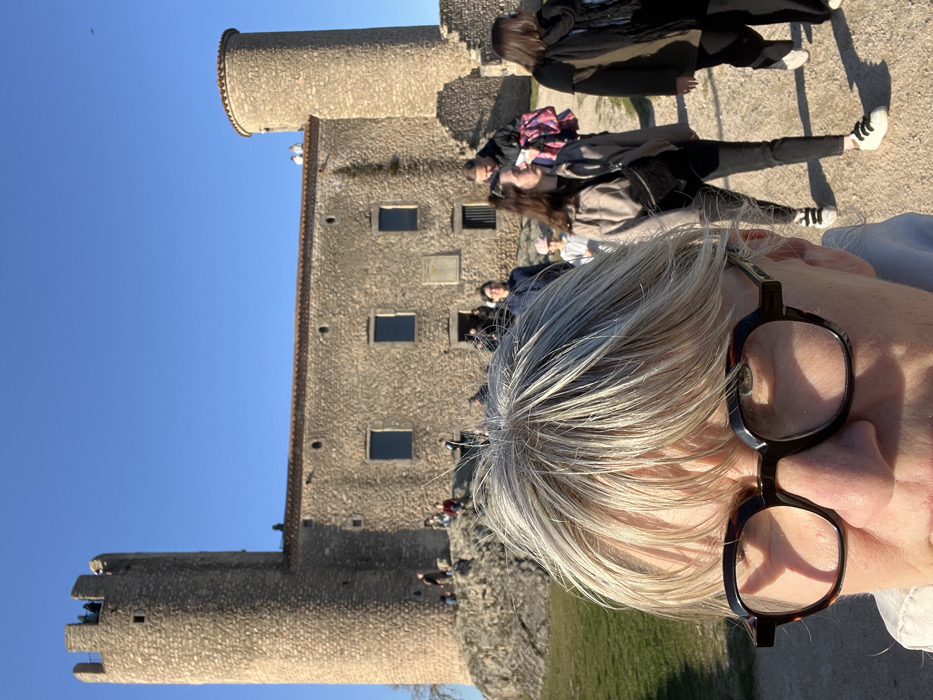 The image depicts an outdoor scene at the Château de Chambles in Chambles, France, under a clear blue sky. In the immediate foreground, the top portion of a person's head is visible, featuring light blonde to grey hair with bangs and dark, square-framed eyeglasses with a tortoise-shell pattern.
The midground shows a dirt path occupied by several pedestrians. A woman with dark hair, dressed in a light beige jacket, dark pants, and white sneakers, walks from left to right, carrying a black sling bag. Behind her, a man with dark hair carries a pink and purple patterned bag. Multiple other individuals are visible walking or standing on the path and surrounding rocky terrain, including a child wearing a pink hat.
The background is dominated by the stone structure of the Château de Chambles. It comprises a central building with a reddish-brown roofline, constructed from rough-hewn light-colored stone, featuring multiple rectangular windows with dark panes. Two prominent round towers flank the central building. The left tower is tall and cylindrical with crenellated battlements, upon which several individuals are standing. The right tower is shorter, also cylindrical, and topped with a rounded, reddish-brown cap. People are also observed on the roofline between the main building and the right tower. The scene is illuminated by strong sunlight.