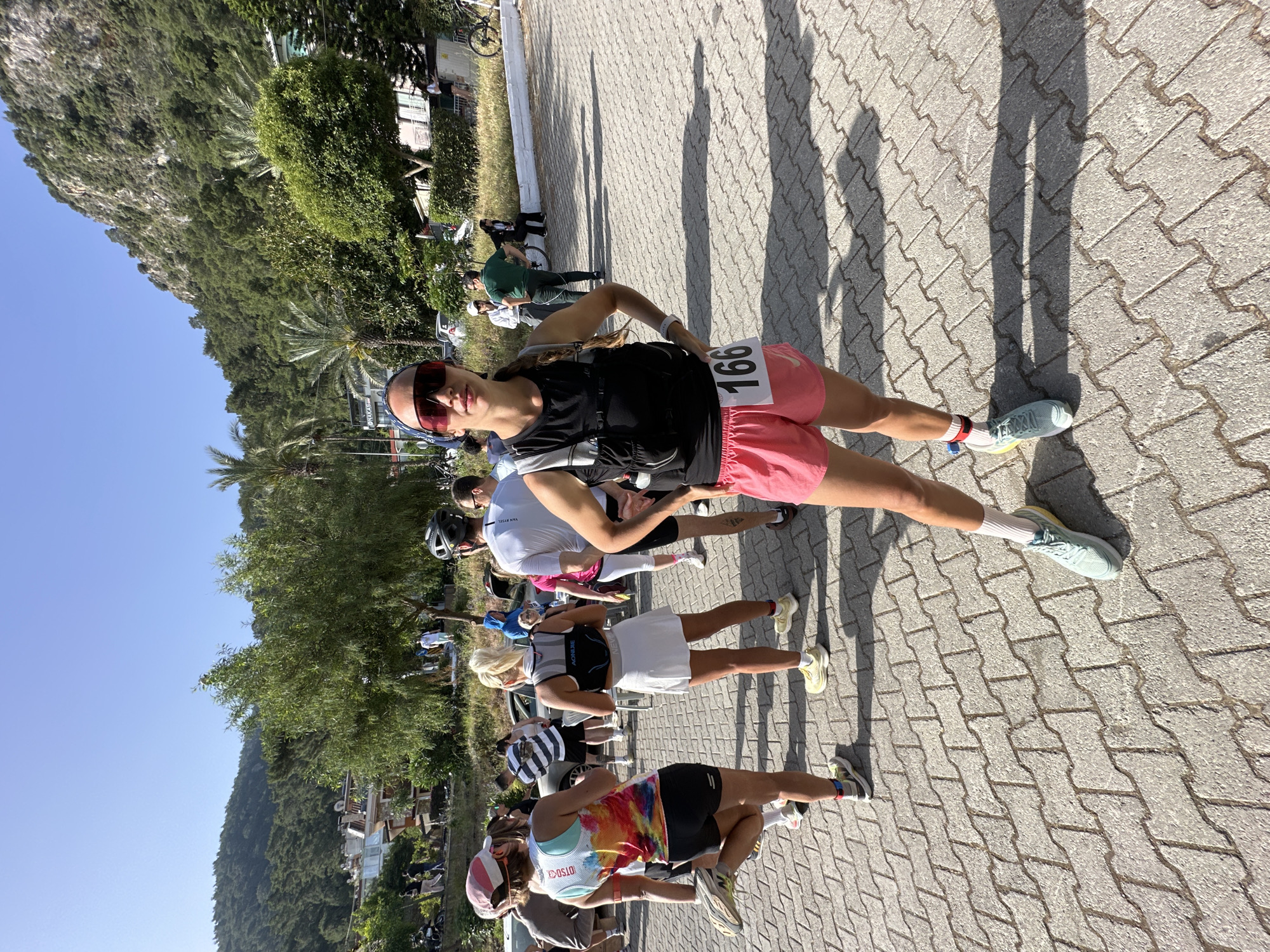 A woman wearing sunglasses and a headband, and sporting the number 166, is standing on a brick-paved road with a group of other people. They are lined up, seemingly waiting for a race or event to begin. The woman is wearing a black shirt and red running shorts. A forested hill rises sharply in the background. This photograph was likely taken in Fethiye, Turkey.