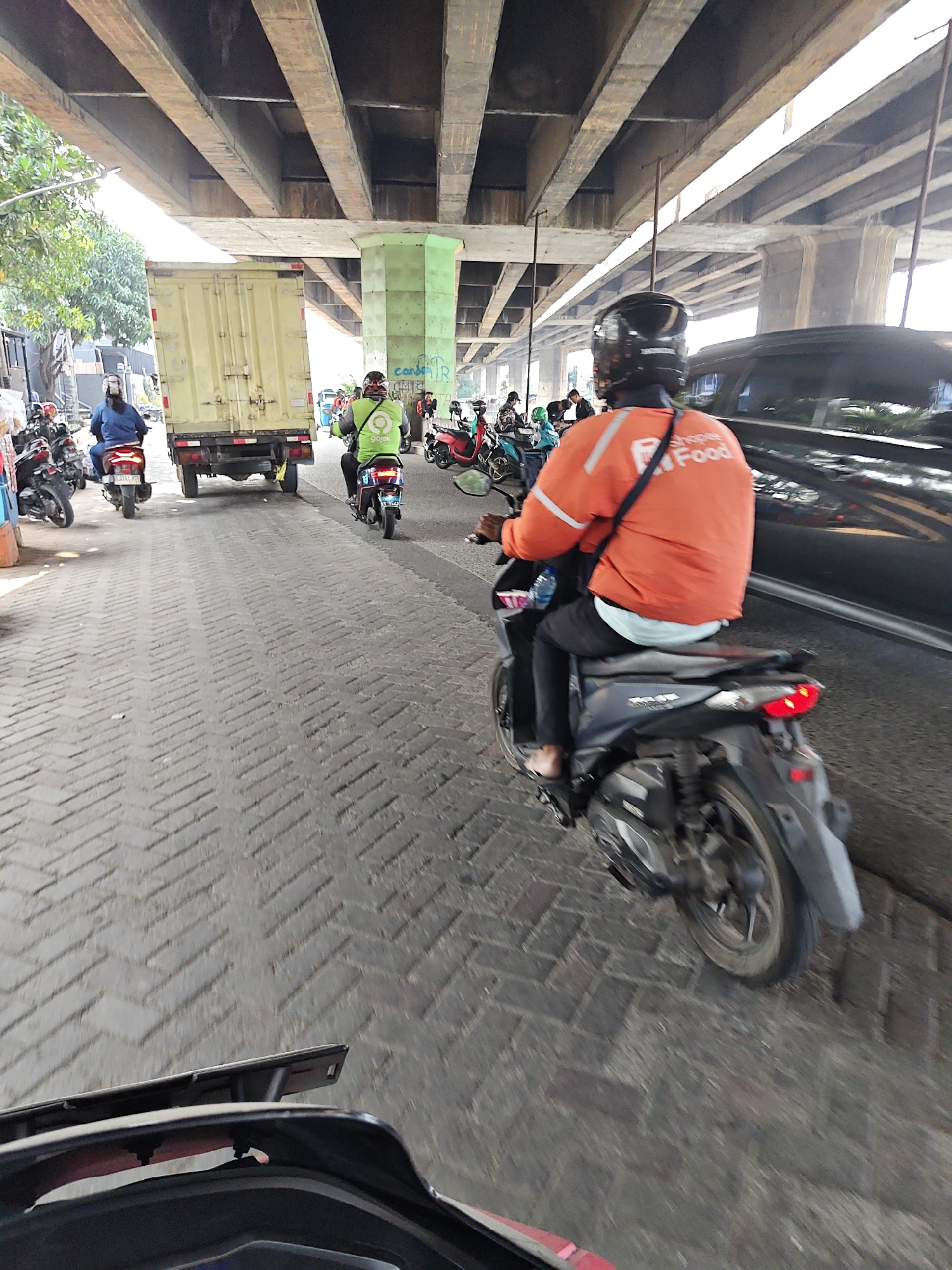 The image shows a busy street scene under a highway overpass in North Jakarta, Indonesia. Several motorbikes are present, with riders wearing helmets and one rider in an orange jacket with "Shopee Food" branding. A yellow truck is parked on the side of the road. The road surface is paved with interlocking bricks. The overpass is constructed with concrete beams and pillars. The overall atmosphere suggests a typical urban environment with significant traffic.