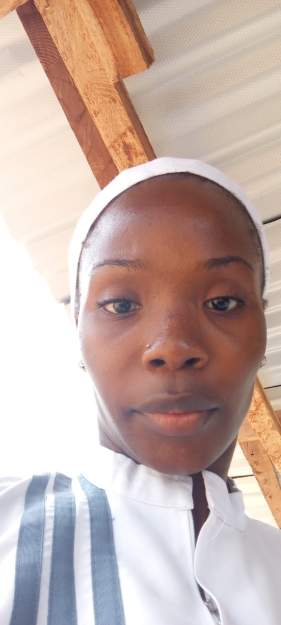 A medium, close-up shot shows a dark-skinned Black woman wearing a white head covering and a white uniform shirt with gray stripes. She has a nose piercing on her left nostril and a small earring on each ear. Overhead are unfinished wooden beams and a white, textured ceiling. The location appears to be indoors, possibly within a building in Keffi, Nigeria.