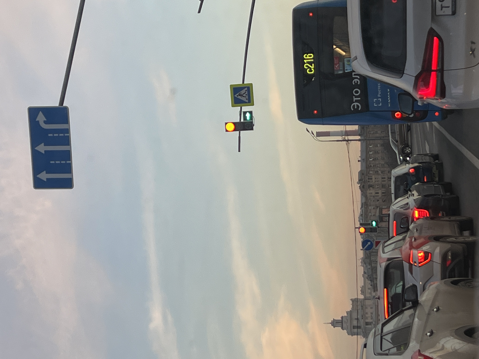 The image depicts a static urban traffic scene on Leningradsky Avenue in Moscow, Russia, with the main building of the Financial University under the Government of the Russian Federation visible on the left in the background. The sky overhead is partially cloudy, showing hues of light blue and soft orange/pink, indicating either early morning or late afternoon.
In the immediate foreground, portions of two vehicles are visible: a light-colored SUV on the left and the rear of a white SUV, possibly a BMW X1, on the right, displaying illuminated red taillights. Further into the midground, a line of stationary vehicles occupies multiple lanes. On the left, a silver/grey SUV, possibly a Hyundai Creta, with illuminated red taillights, is followed by other cars. In the adjacent lane on the right, a blue city bus, identified as a Kamaz vehicle with "Ростех" branding and "Это ЭЛ" in Cyrillic, displays the route number "c216" in yellow LED characters on its rear, along with illuminated red taillights. Another white SUV with illuminated red taillights is directly behind the bus.
Above the street, a blue rectangular overhead sign with white arrows indicates lane usage: two straight-through lanes and one right-turn lane. To the right of this sign, a vertical traffic light shows a solid red circular signal for vehicular traffic, with a smaller, horizontal green arrow pointing right illuminated below it, indicating a permissive right turn. Adjacent to the traffic light, a square blue pedestrian crossing sign with a white walking figure is mounted. In the far background, behind the bus, a red and white striped industrial chimney is visible. Vehicles are stationary, indicating traffic congestion at a red light. No individuals are clearly visible.