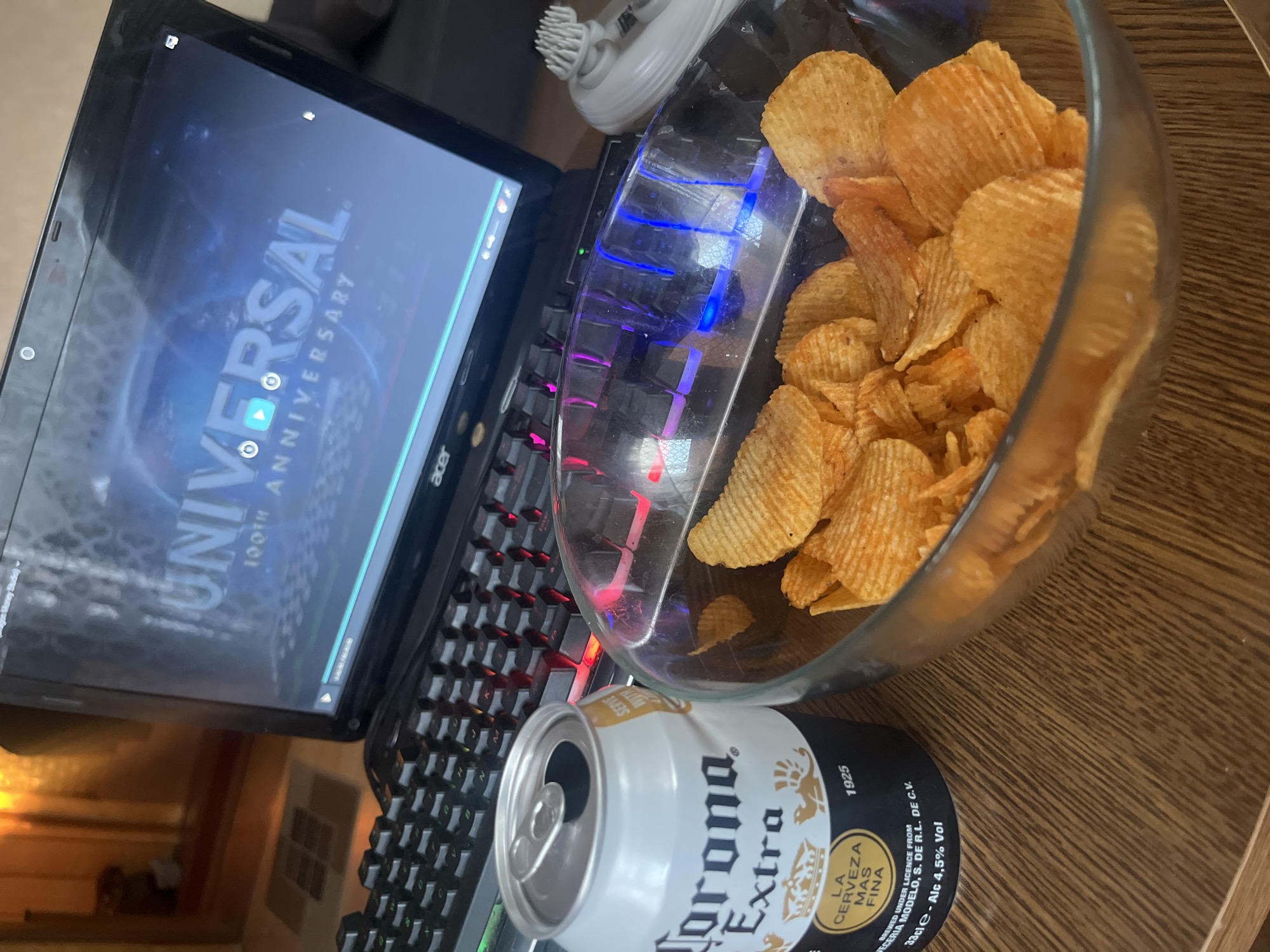 The image depicts a casual home setting, likely a desk or table, with a laptop open and displaying the Universal Pictures logo with "100TH ANNIVERSARY" text. In front of the laptop is a keyboard with illuminated keys, suggesting it might be dark or the backlighting is on. To the left of the keyboard is a can of Corona Extra beer, and a glass bowl filled with ruffled potato chips sits in front of the keyboard. To the right of the bowl, a small white electronic device, possibly a facial cleansing brush or massager, is visible. The overall scene conveys a moment of relaxation, with someone likely watching a movie or video while enjoying snacks and a drink. The image does not contain any visual cues or information that would indicate a specific location in Białystok, Poland.