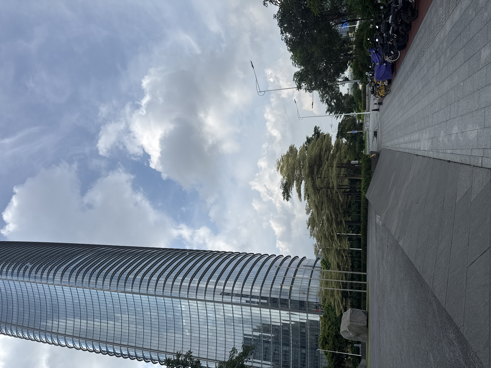 A full-shot, low-angle, outdoor photograph captures a modern cityscape dominated by a tall, curved skyscraper with a glass facade. The building occupies the left side of the frame, its reflective windows mirroring the cloudy sky. In the foreground, a paved area transitions into a sloped, dark grey textured surface. To the right of the skyscraper, a line of trees with light-colored foliage forms a visual barrier. Beyond the trees, a street is visible, lined with streetlights and parked motorcycles, some covered with purple tarps. A large, weathered rock is situated on a patch of grass in the mid-ground. The sky is filled with a mix of white and grey clouds against a pale blue backdrop. The scene appears to be in Haizhu District, China.