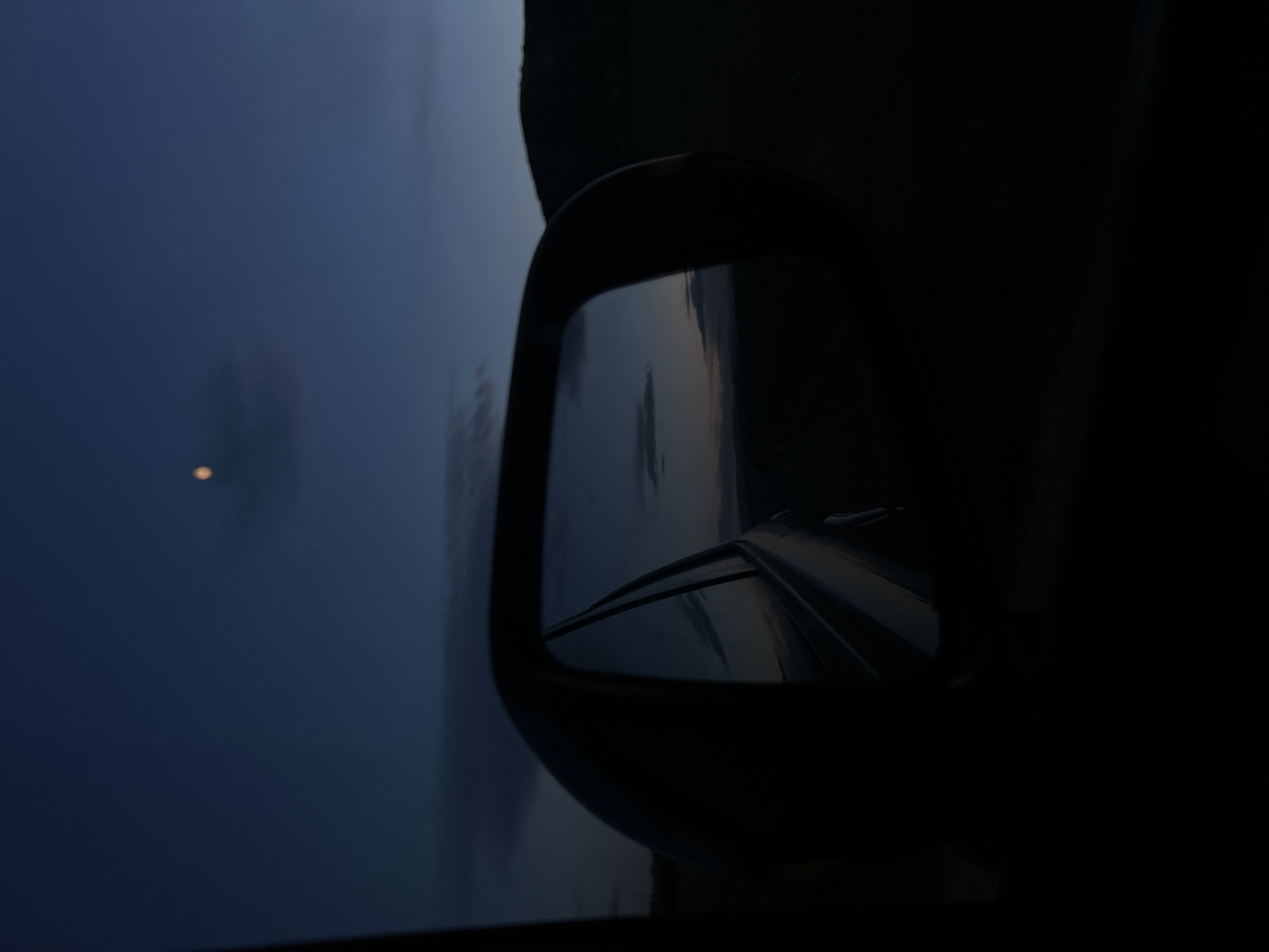 The image depicts a car side mirror reflecting a road and sky at twilight or dawn. The sky is a dark blue with a possible moon or bright star visible. The road is also dimly lit, with a faint reflection of the sky visible on its surface. The side mirror is in the foreground, and the rest of the car interior is obscured by darkness. The image does not provide any indication of the location being in the city of Ovchepoltsi, Bulgaria.