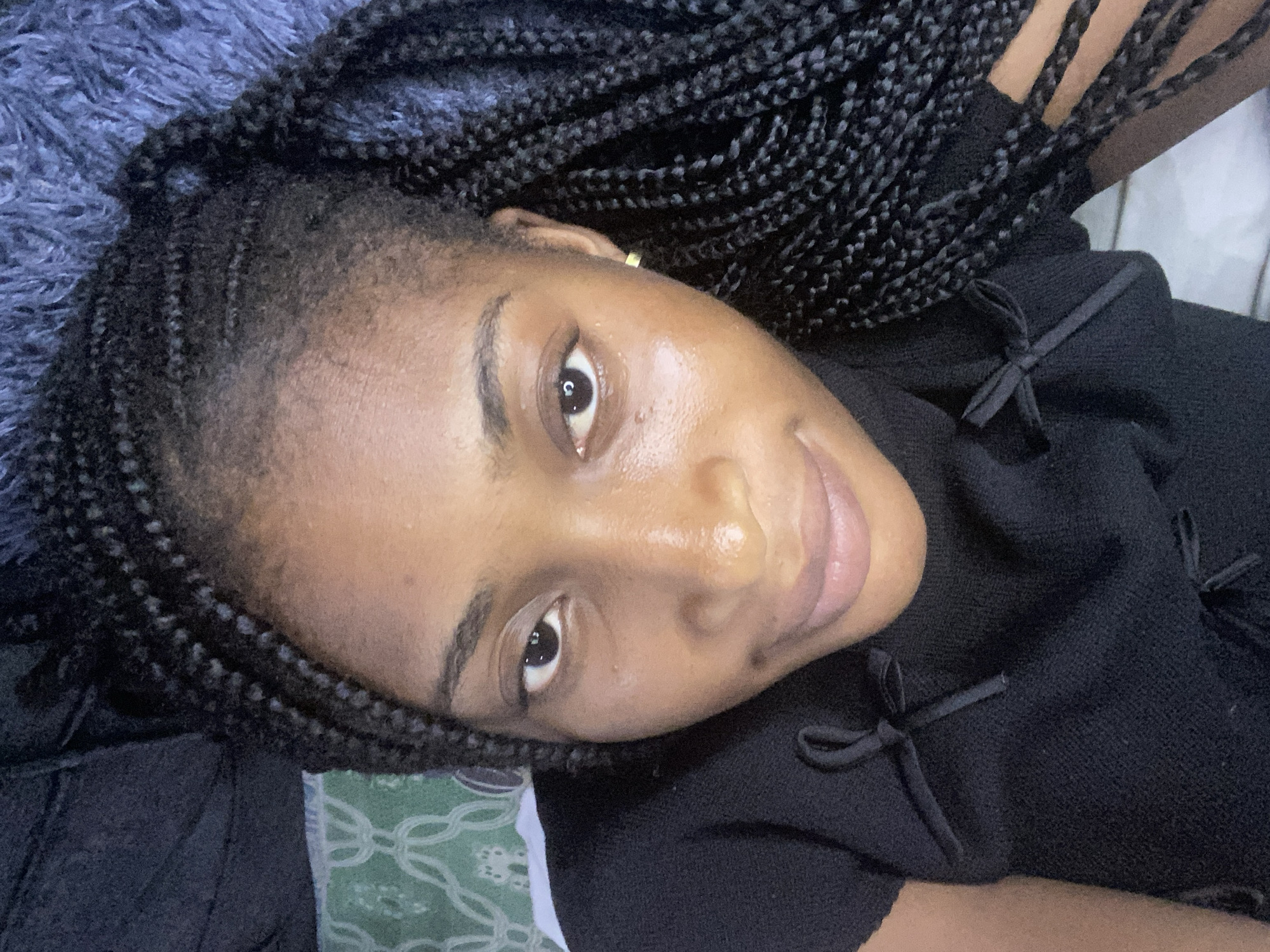 A young Black woman with braided hair is lying down, looking up at the camera. She is wearing a black top with a high neckline and two bow details on the front. The background appears to be a bed with a textured blue throw blanket and a patterned pillow. The lighting suggests an indoor setting, possibly a bedroom in Kubwa, Nigeria.