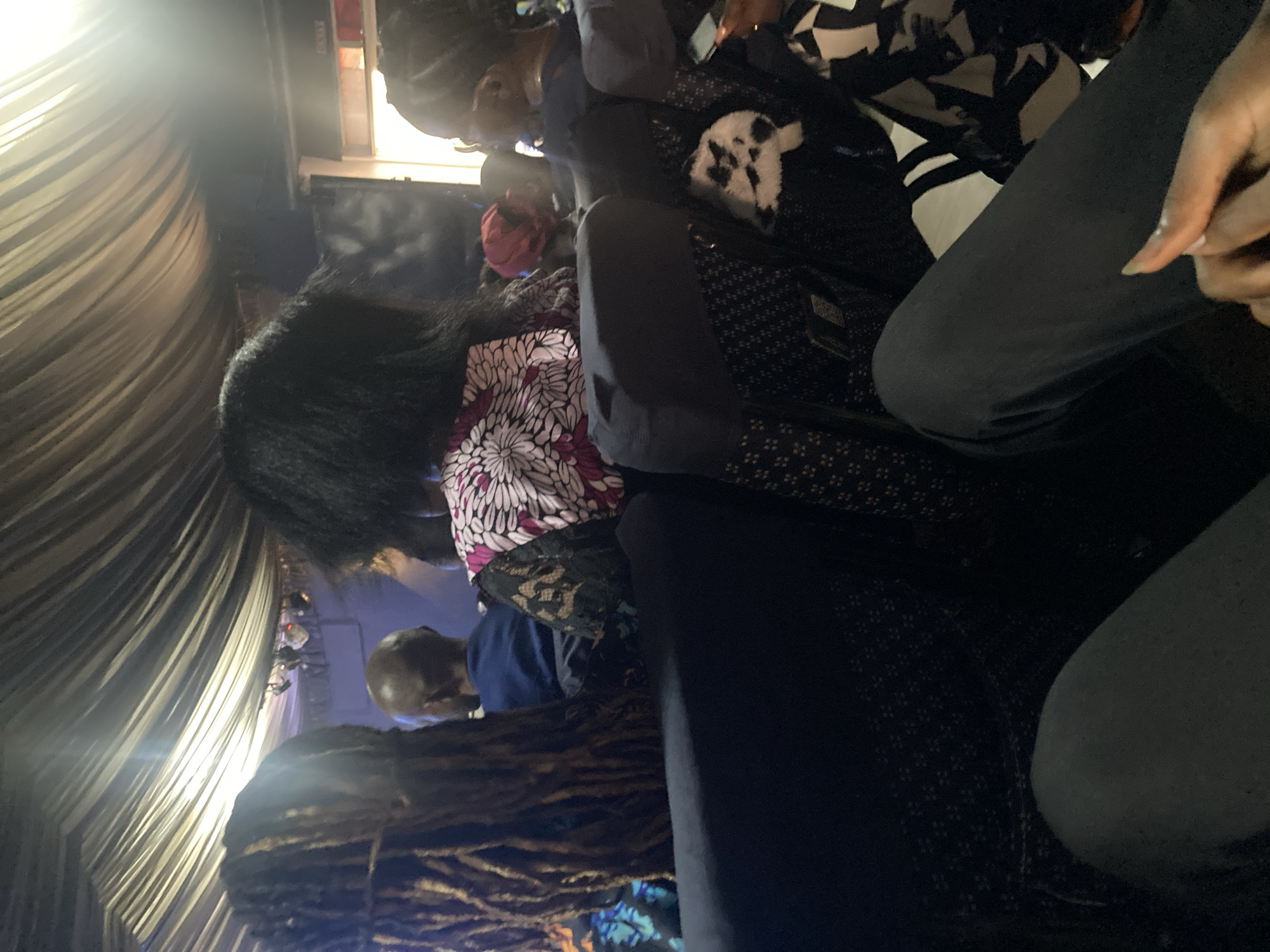 The image appears to be taken inside a tent or covered area, possibly in the city of Jos, Nigeria. Several individuals are visible, seated in what seems to be a crowded space. One person has long dark hair and wears a top with a white and pink floral pattern. Another individual is wearing a dark top with a patterned design. There is a person with short hair or a shaved head wearing a blue shirt. Some sort of dark, textured material is in the foreground of the image. Light is entering from the left side, casting a glow on the tent fabric.