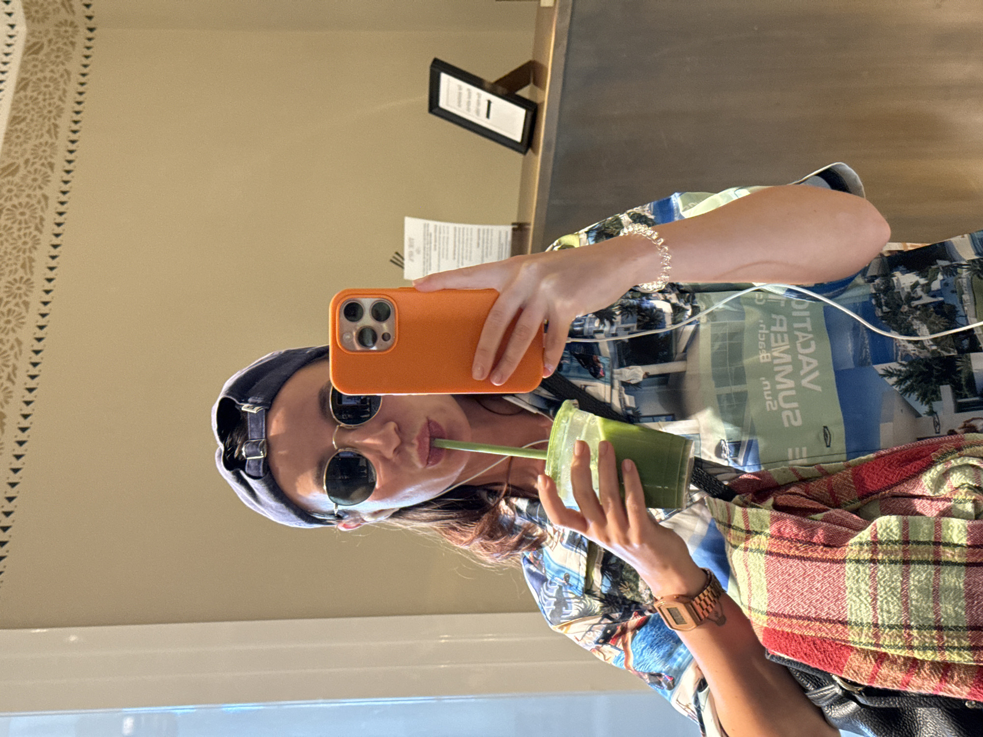 A woman is taking a selfie in a brightly lit room, possibly a cafe or hotel lobby. She is wearing a patterned shirt, a blue baseball cap, and round sunglasses. She is holding a green drink with a straw in her left hand and an orange smartphone in her right hand, which she is using to take the picture. The background shows a wall with decorative trim and a counter with some signs and a framed picture. The image has a casual and personal feel, suggesting a candid moment captured by the woman herself. The room's decor appears to be modern and clean, with warm lighting.