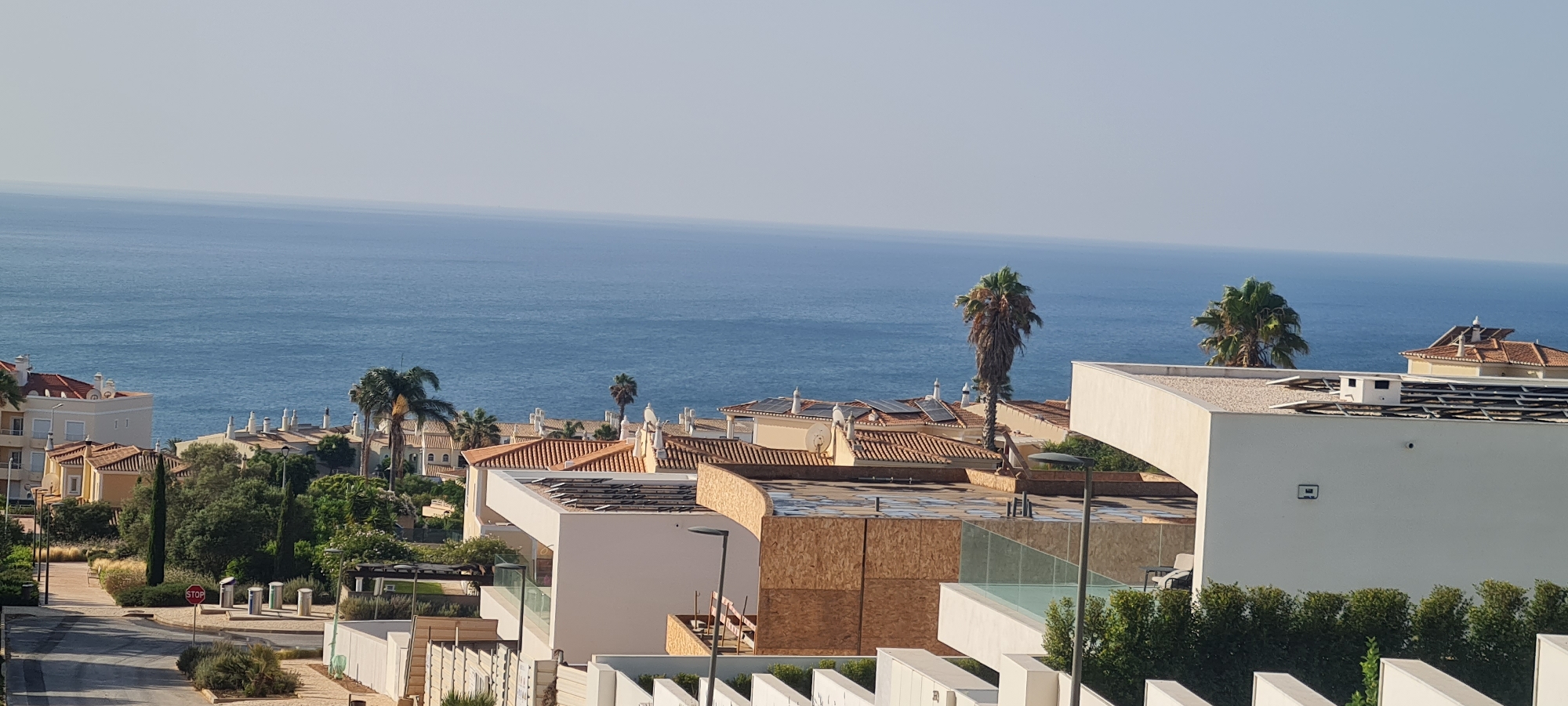 This is a high-angle, wide shot of a coastal residential area, likely in Lagos, Portugal, as suggested by the architectural style and lush vegetation. The foreground shows a street sloping downwards, lined with modern white fences and manicured green bushes. A red stop sign is visible on the side of the street, along with several recycling bins.
Dominating the midground are several multi-story buildings with terracotta tile roofs. Some of these buildings appear to be apartment complexes or villas, with light-colored facades and white window frames. One prominent building in the mid-right is under construction, with exposed wooden framing on its exterior and a modern white facade with glass railings. Palm trees of varying sizes are scattered throughout the residential area, with one particularly tall, slender palm tree standing out in the mid-right.
In the background, the deep blue ocean stretches to the horizon, meeting a pale, hazy sky. The sea appears calm with very small waves. The overall impression is a sunny, Mediterranean-style resort or residential neighborhood overlooking the sea. There are no people visible in the image.
