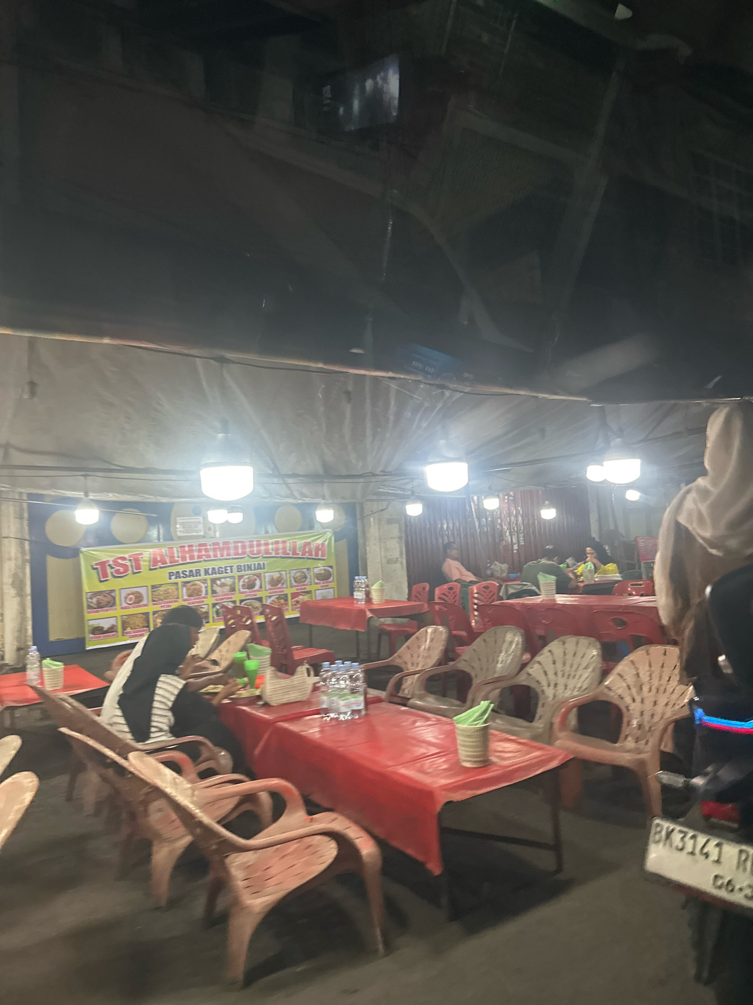 An outdoor food stall area in the city of Binjai, Indonesia, is depicted at night. The scene is illuminated by overhead lights and a banner displaying "TST ALHAMDULILLAH PASAR KAGET BINJAI" with images of food items. Red plastic tables covered with red cloths are set up, accompanied by plastic chairs of various colors including red, beige, and white. Several people are seated at the tables, some appearing to be dining or socializing. A motorcycle with a license plate reading "BK3141 RB" is parked on the right side of the frame. The overall environment suggests a bustling street food market.