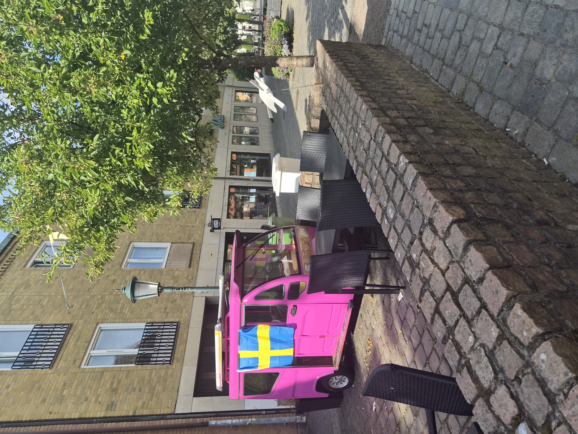 In Varberg, Sweden, a bright pink mobile vendor cart, adorned with a Swedish flag, is parked on a cobblestone street. The cart appears to be a food or beverage service, with a serving window visible. Outdoor seating, consisting of dark-colored chairs and tables, is set up on the cobblestone area in front of a low brick wall. In the background, a tree with lush green foliage provides shade, and shops with storefronts line the street. The scene is illuminated by natural daylight, suggesting it is daytime.