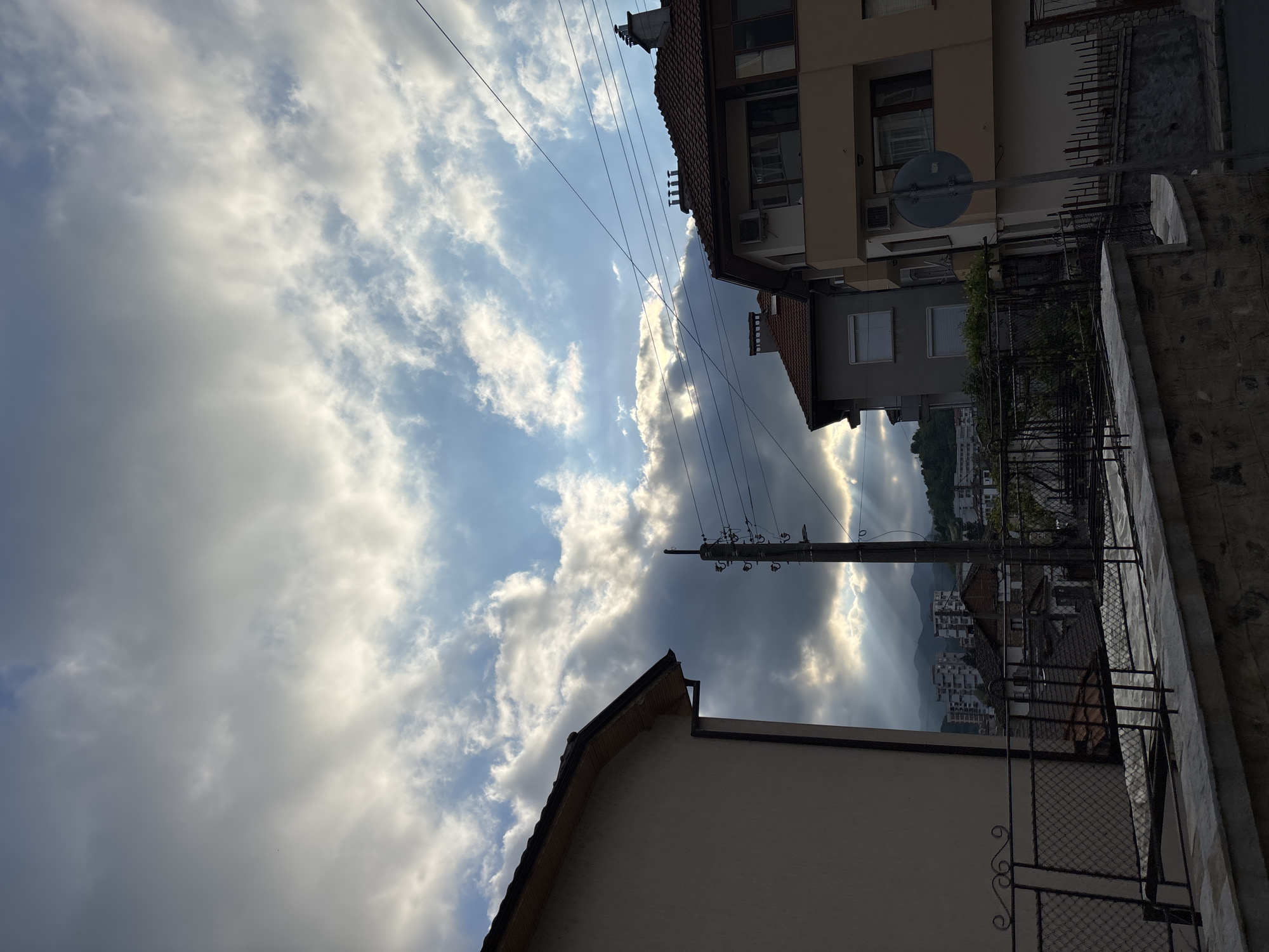 The photograph depicts a partly cloudy sky above a residential area in Zlatograd, Bulgaria. The view is upward from street level, showing the tops of buildings and an electrical pole with wires against the backdrop of clouds and blue sky. Rays of sunlight break through the clouds, illuminating the distant hills and buildings in the valley. A portion of a building with a red tile roof is visible in the foreground, and the side of another building is seen adjacent to a fence with decorative ironwork. The composition is vertical, with the sky occupying a significant portion of the frame.