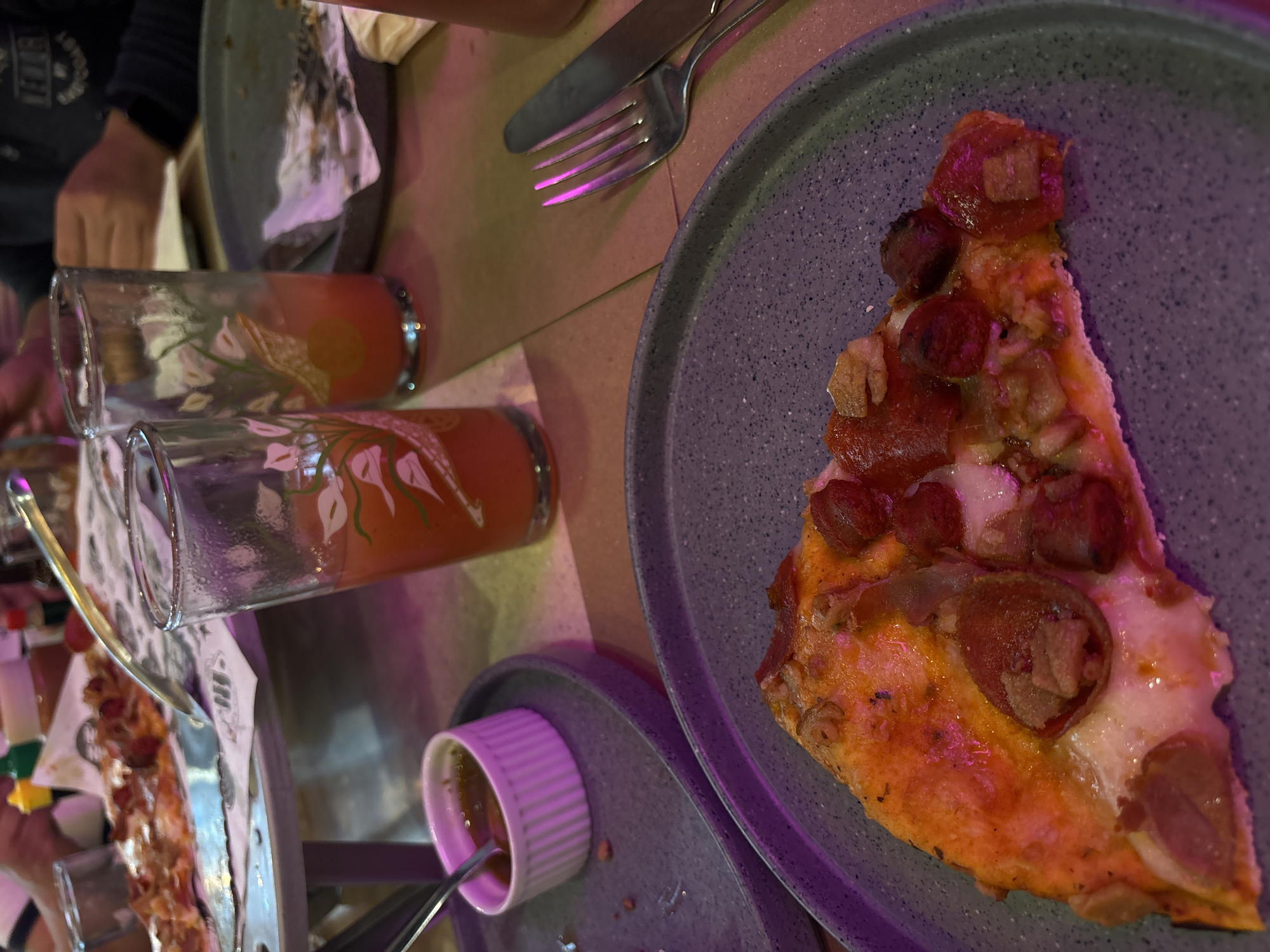 The image shows a tabletop scene at a restaurant, likely in Puebla, Mexico, based on the overall aesthetic and common restaurant setups. Present are two glasses filled with orange-colored liquid, potentially juice or a cocktail, lying on their sides. A light gray plate holds a single slice of pizza, topped with sausage or pepperoni, cheese, and what appear to be small pieces of onions. Adjacent to the pizza plate is a smaller light gray saucer containing a small, white ramekin with a dipping sauce and a spoon inside. A fork and knife rest on the table. A portion of another plate with food is partially visible on the left. A person's arm and hand can be seen on the left side of the frame. The tabletop appears to be textured or made of individual tiles or sections. The lighting is subdued with an artificial glow, indicating an indoor setting.